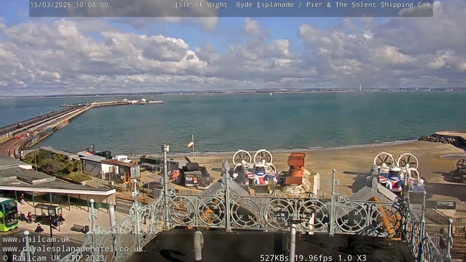 A partly cloudy day shows a long pier extending into a calm body of water, with hovercrafts parked on a sandy shore nearby.
