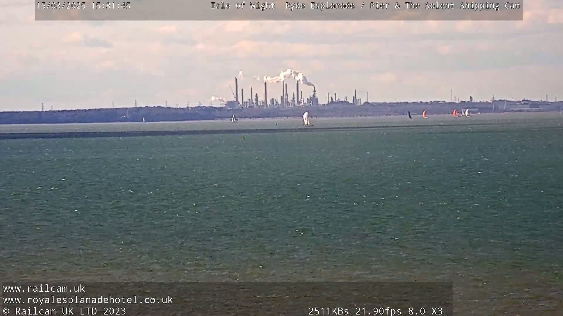 A body of water with several sailboats is seen in the foreground, with an industrial plant emitting smoke visible in the distance under a mostly clear sky.