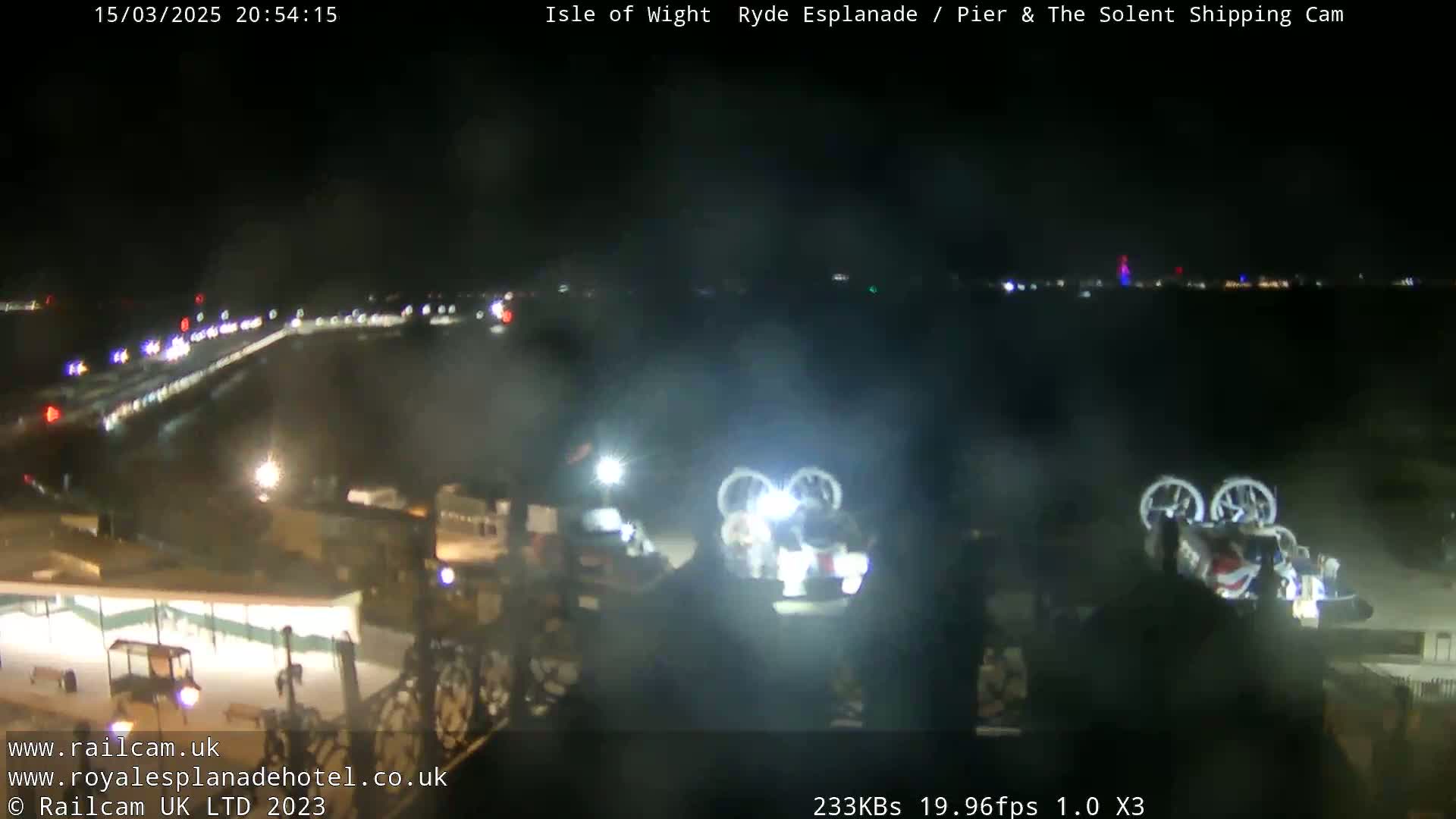 A nighttime view of a waterfront area with two hovercrafts illuminated near a pier, under hazy conditions.