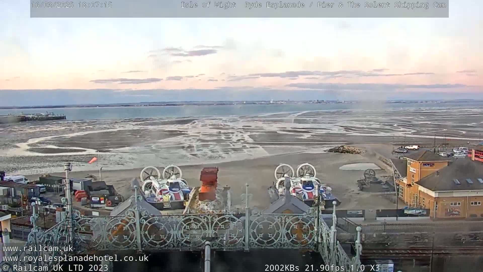 Two hovercrafts are parked near a building on a beach with exposed mudflats and calm water in the background under a partly cloudy sky.
