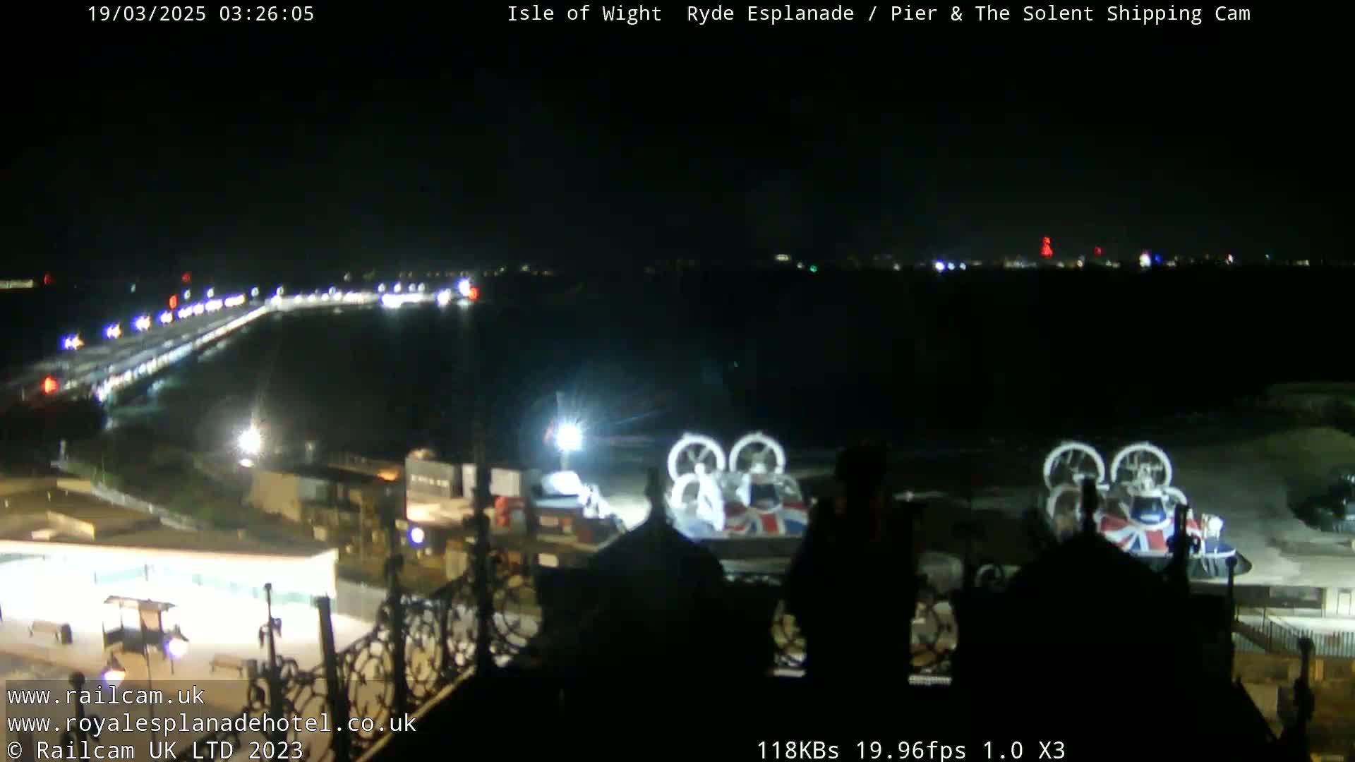 A nighttime, long shot of a pier with lights, a body of dark water, and two hovercrafts parked near a building.