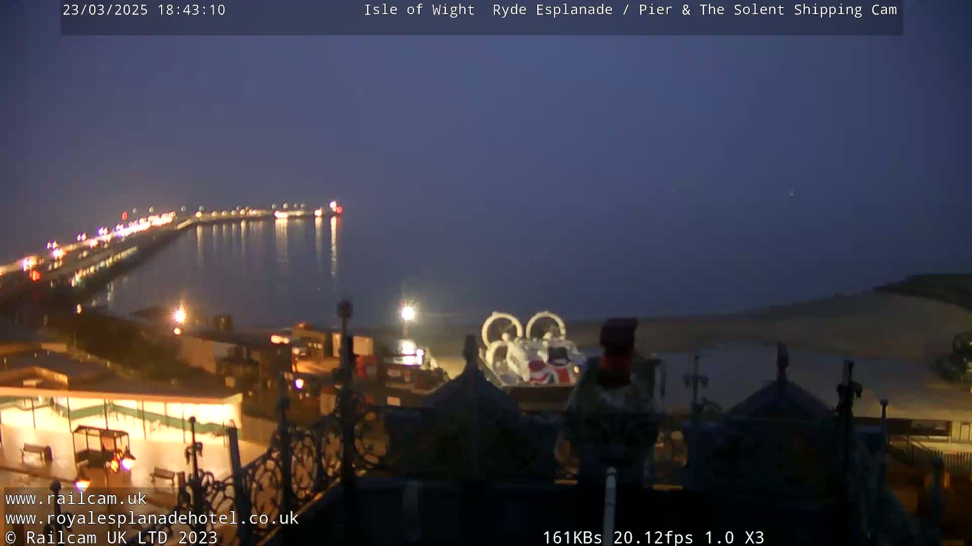 A nighttime view of a pier and calm body of water, with a hovercraft visible on the shore under a dusky sky.