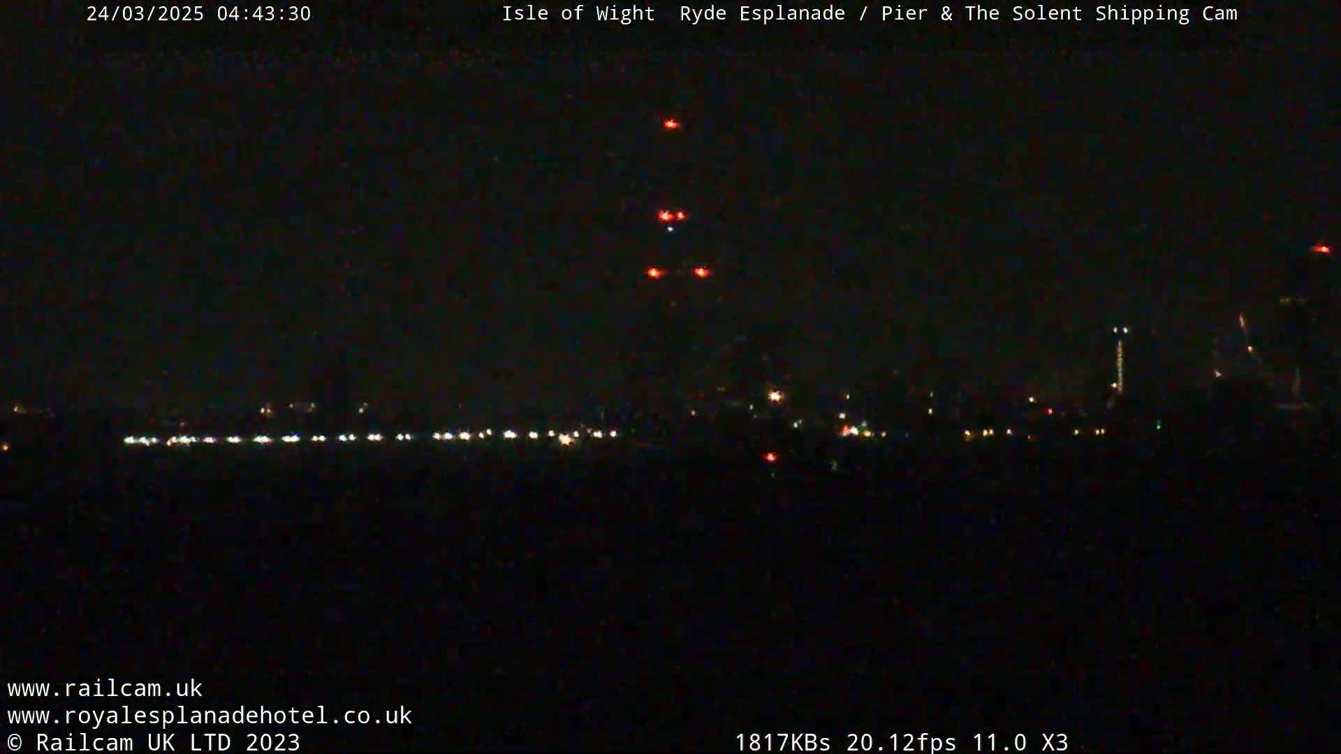 A nighttime, long-range view shows a distant cityscape with a few bright lights and a cluster of red lights in the center, possibly a tower or structure.