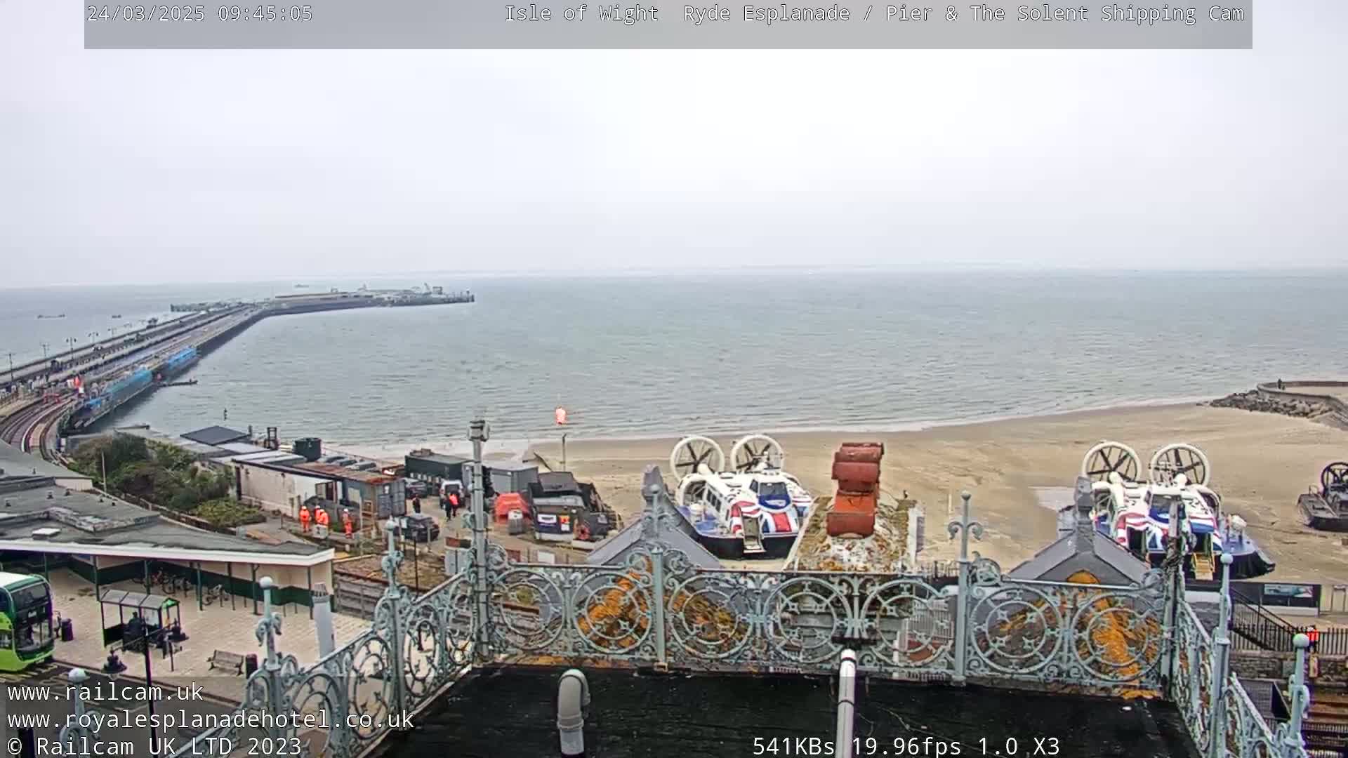 A view from above shows a beach with two hovercrafts, a long pier extending into a calm, gray sea under an overcast sky.