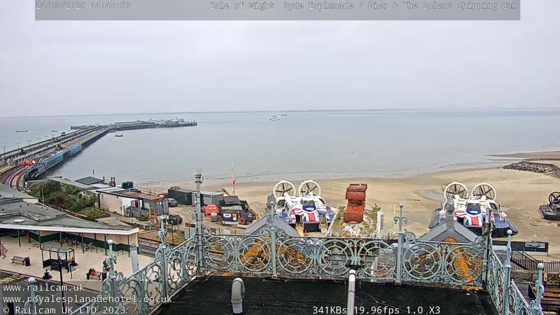 A long pier extends into a calm, gray-sky sea, with hovercrafts parked on a sandy beach nearby.