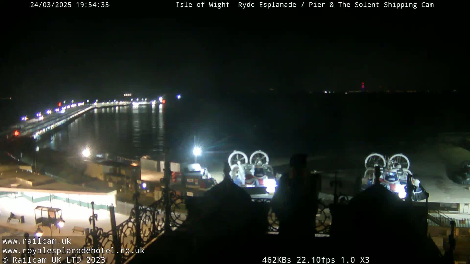 At night, a pier extends into dark water with lights illuminating it, and two hovercrafts are visible on the shore.