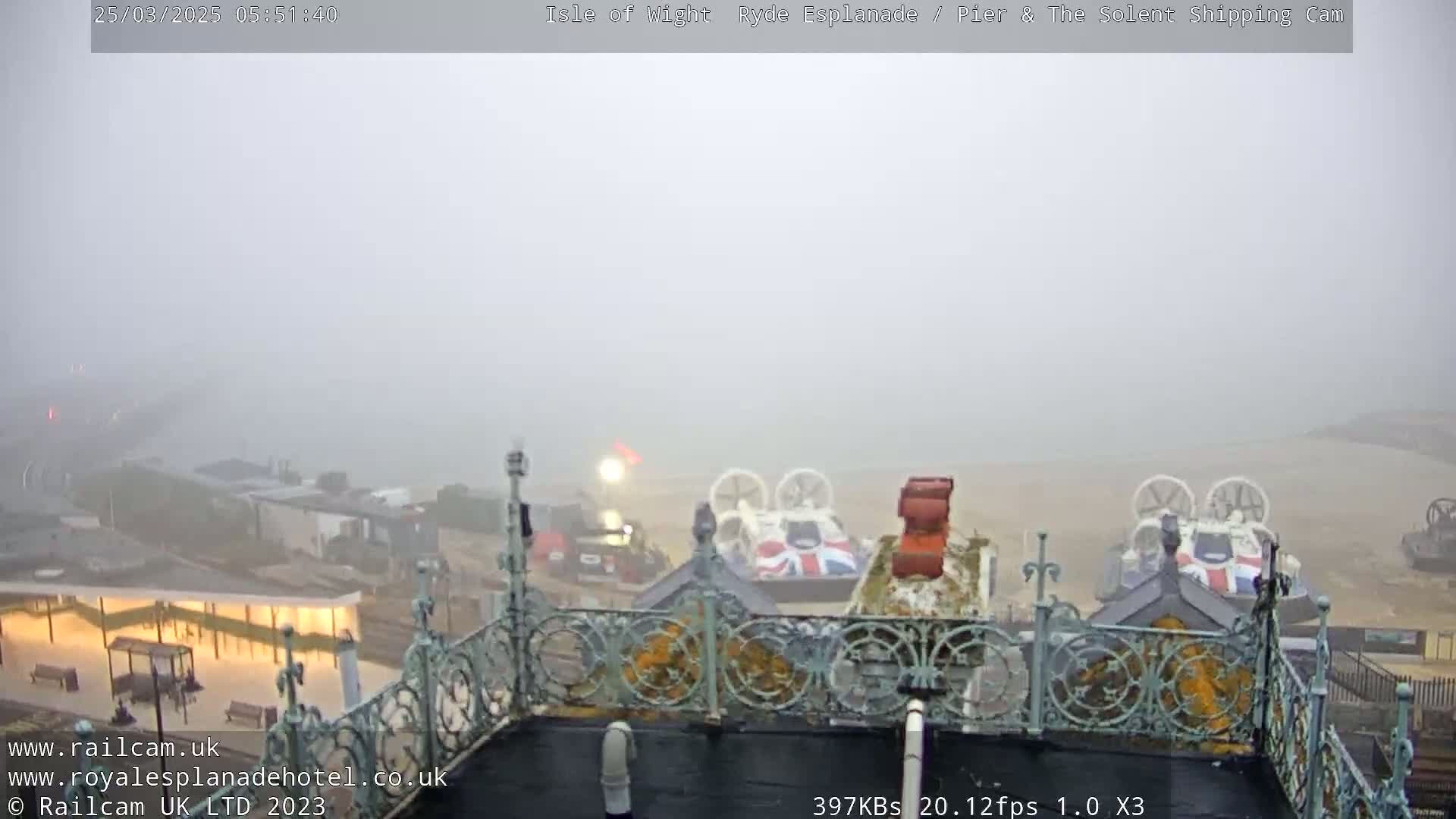 A foggy view from an elevated position shows hovercrafts on a beach near a pier and buildings.