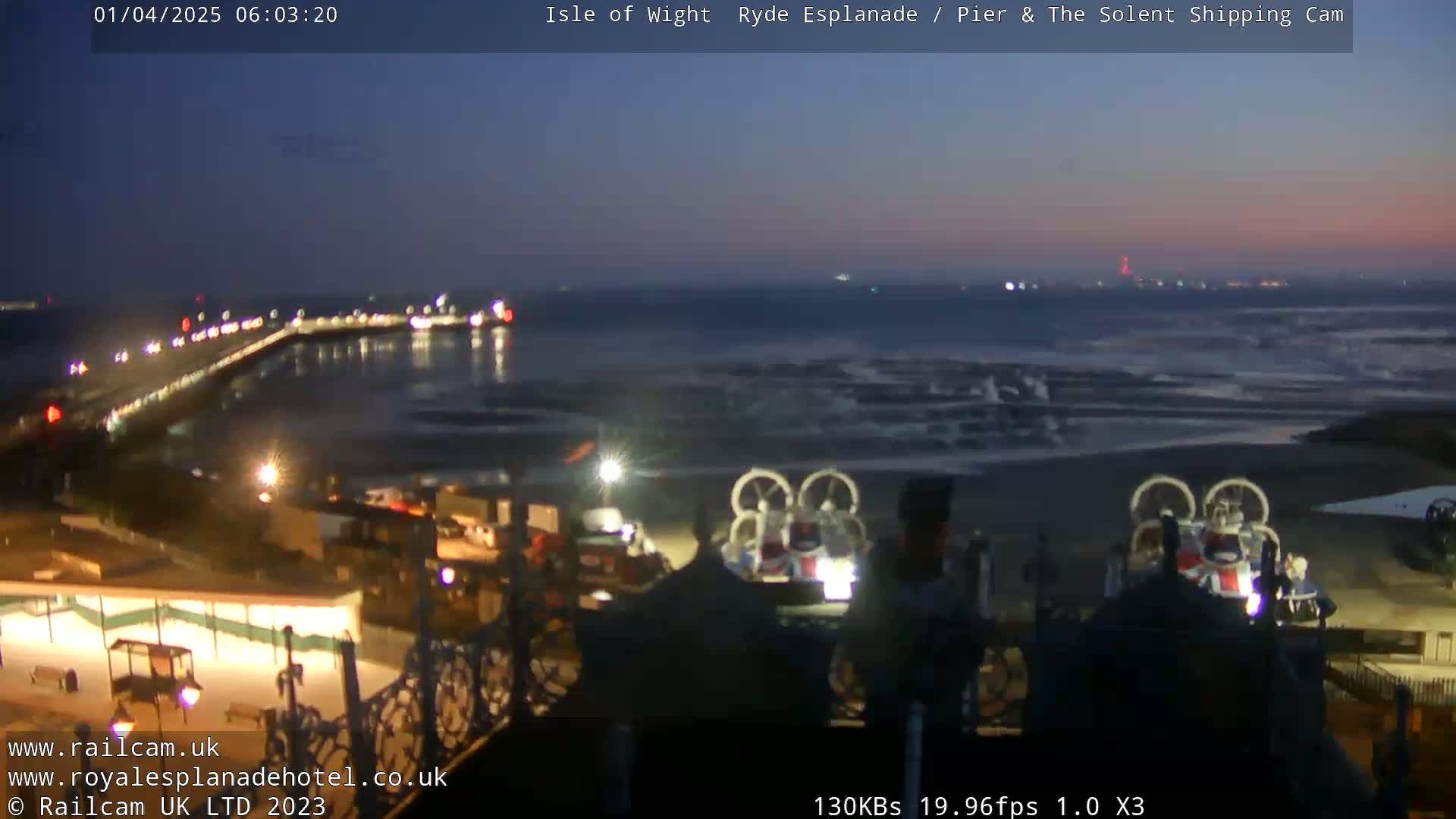 A nighttime view of a pier with lights, a beach with waves, and two hovercrafts on the shore under a twilight sky.