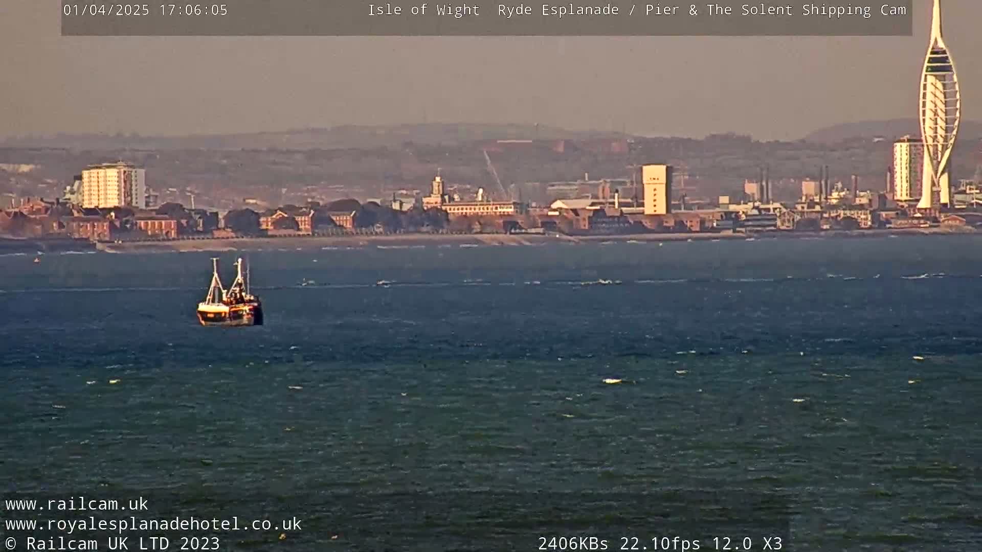 A small fishing vessel is on a somewhat choppy body of water in front of a distant cityscape under a hazy, late afternoon sky.