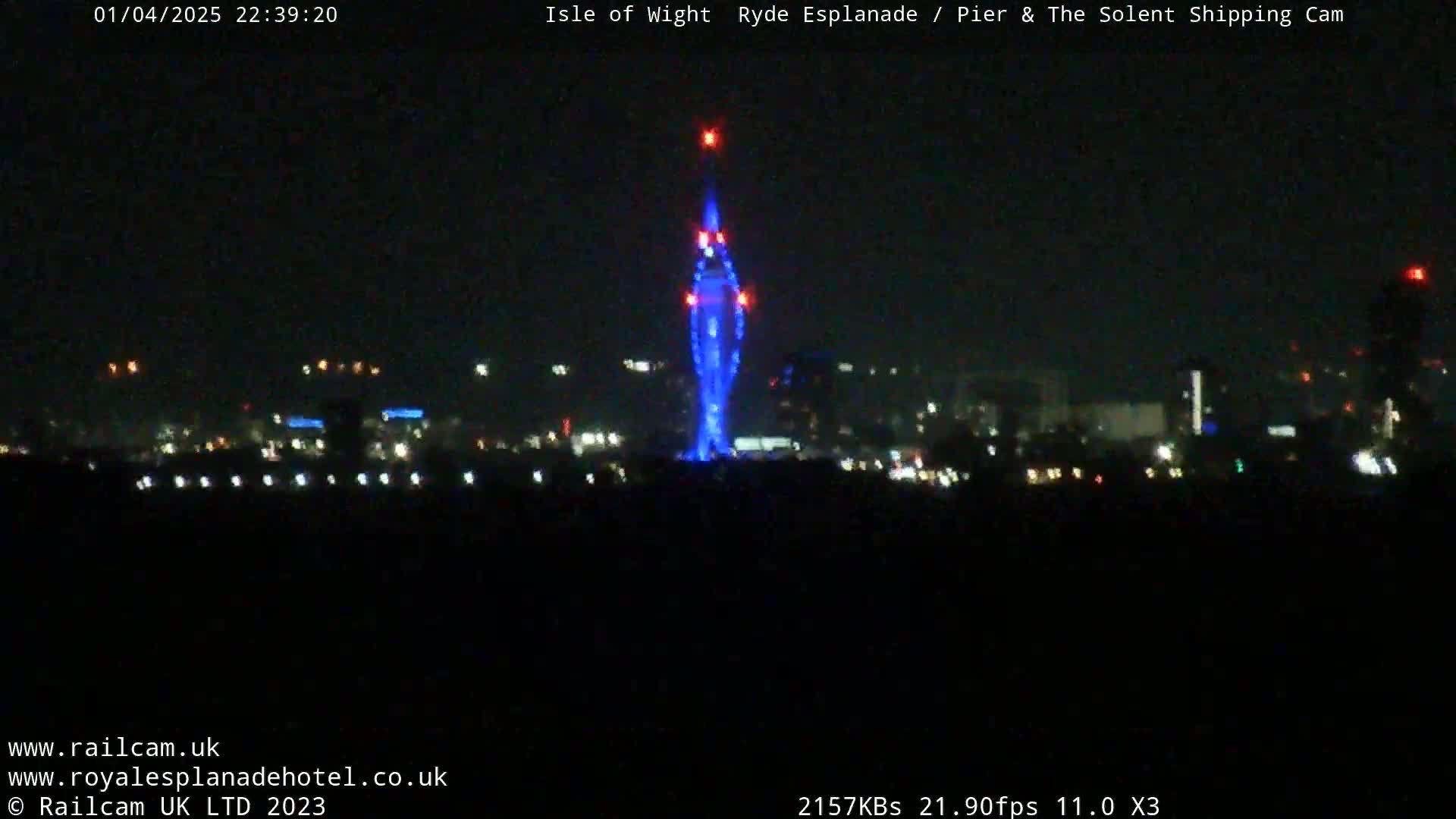 A tall, slender structure is illuminated in blue at night, with a city skyline and dark water visible in the background under a clear, dark sky.