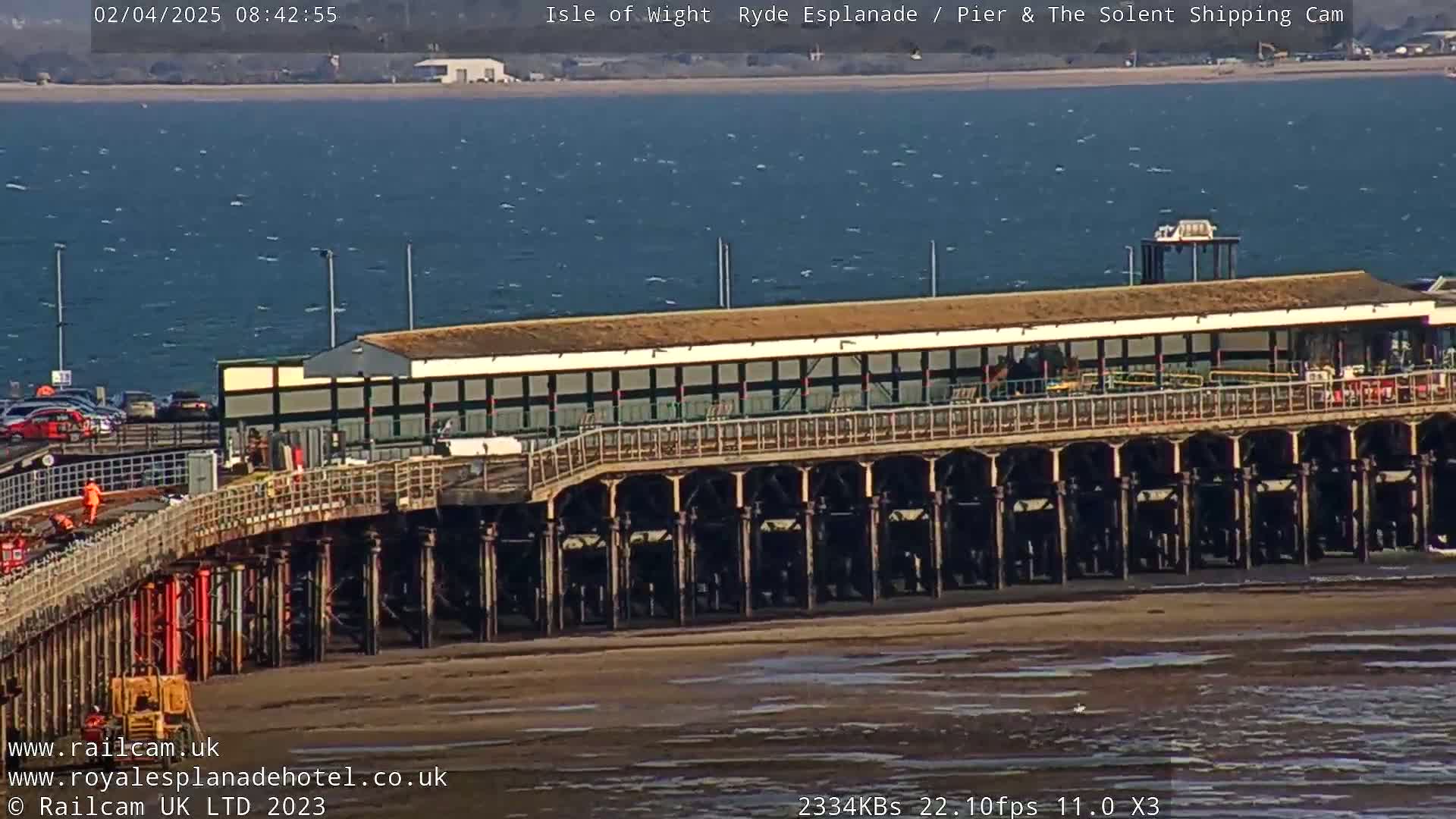 A long pier extends over a calm, slightly windy body of water, with a sandy beach visible in the foreground.