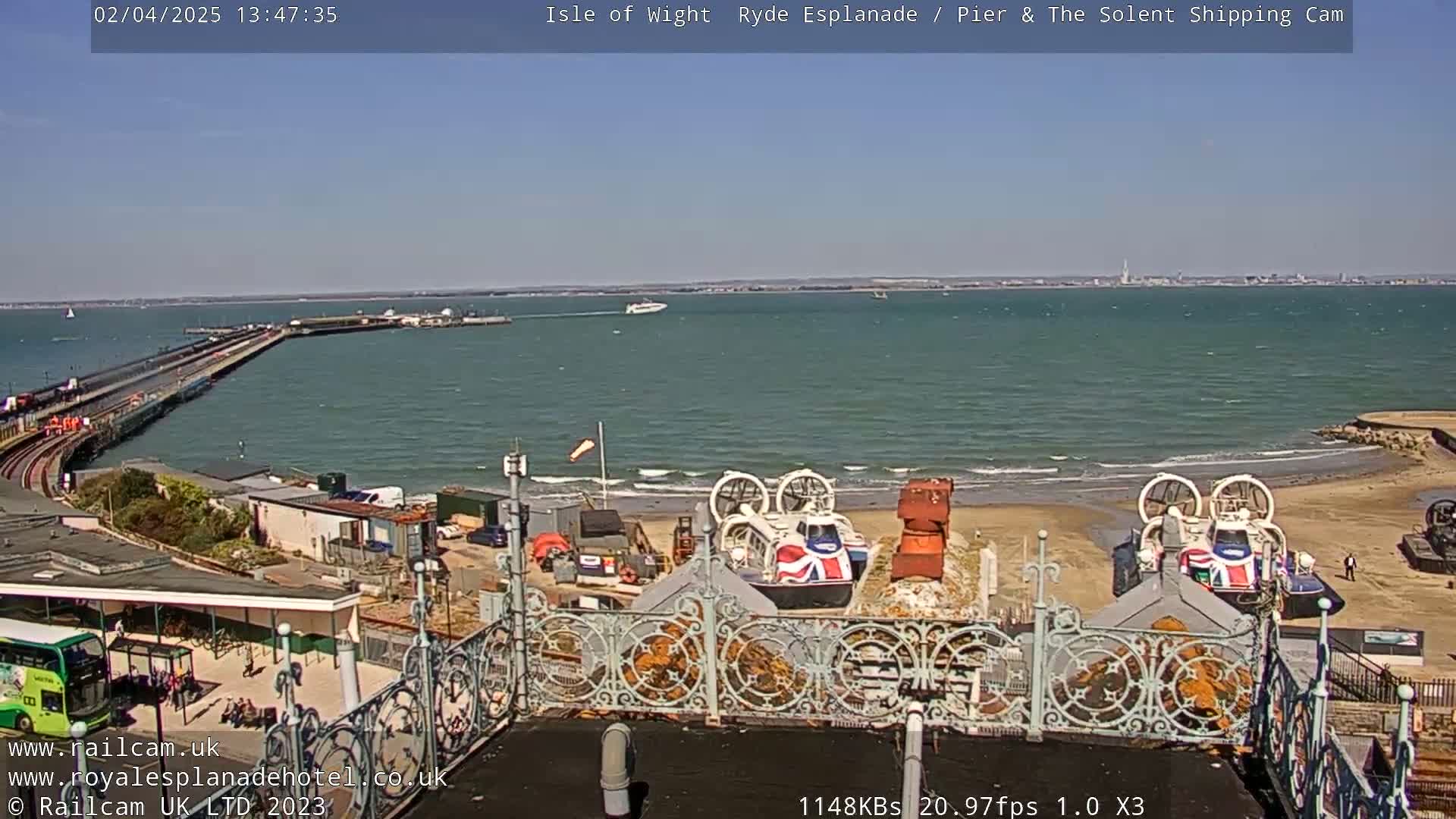 A sunny day shows a pier extending into a body of water with two hovercrafts on a sandy beach, and a distant coastline visible in the background.