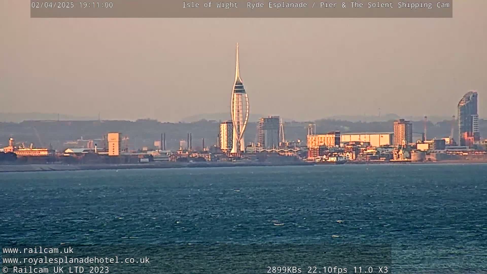 A calm body of water shows a distant cityscape at sunset, featuring a slender, spire-like tower.