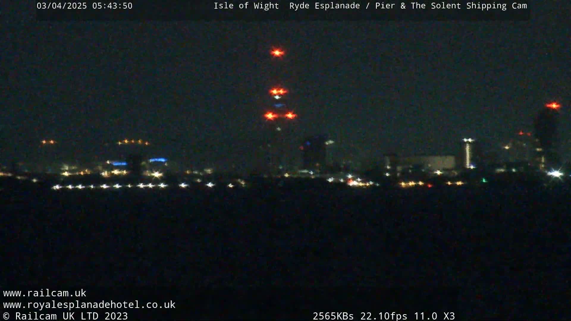A nighttime, long-range view of a city skyline with illuminated buildings and a tall structure with red lights at its top.