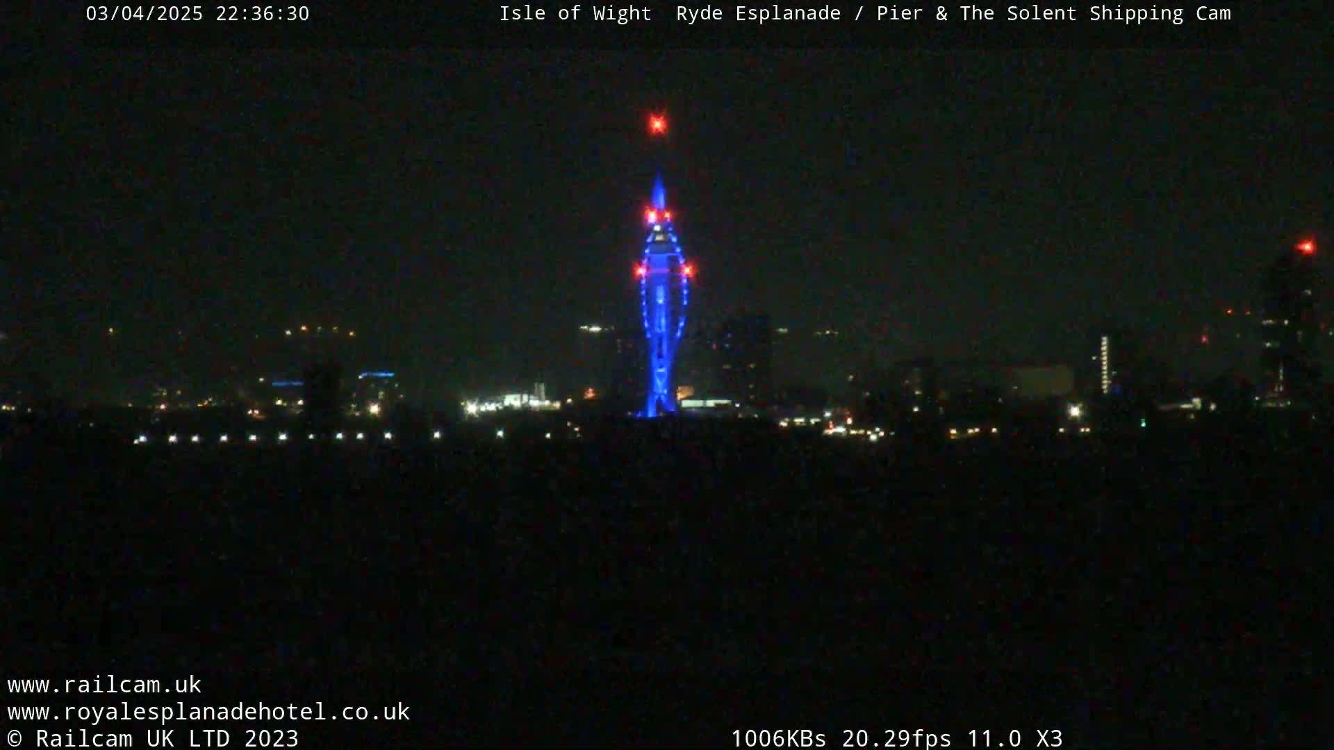 A tall, slender structure illuminated in blue stands out against a dark, nighttime cityscape under a clear sky.