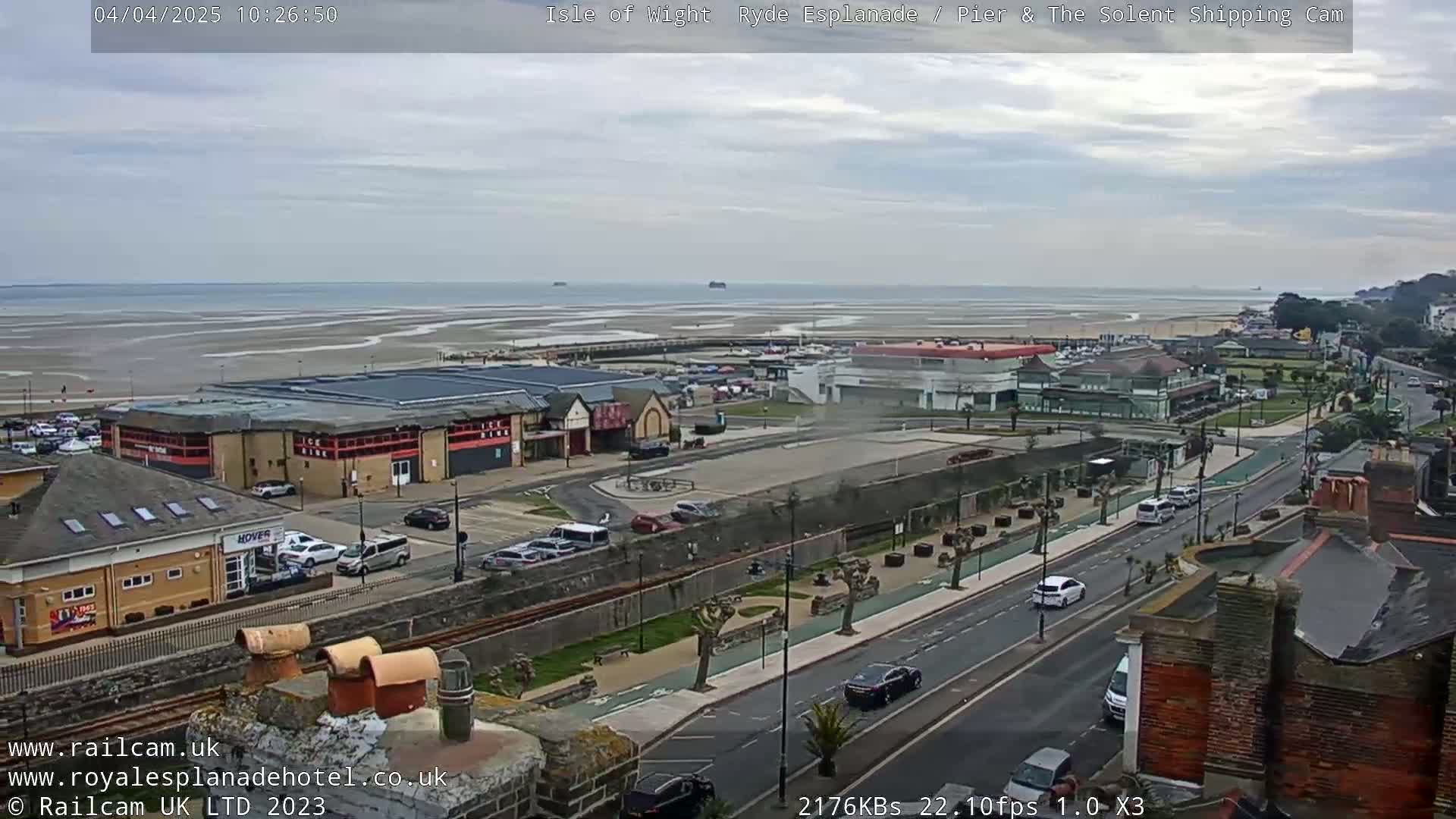 A coastal town scene on an overcast day shows a beach with exposed sand at low tide, buildings along a road, and cars in traffic.
