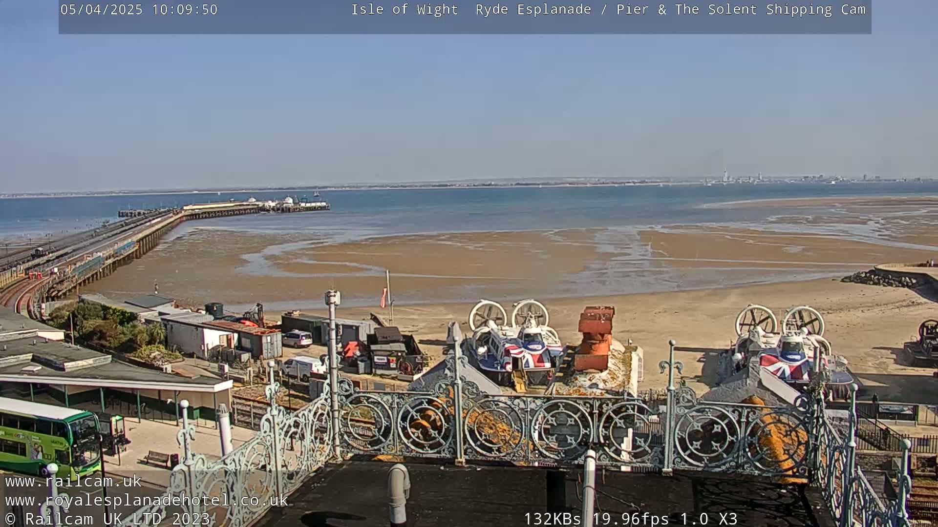 A sunny day shows a pier extending into a wide expanse of calm water, a sandy beach, and two hovercrafts parked near an ornate railing.