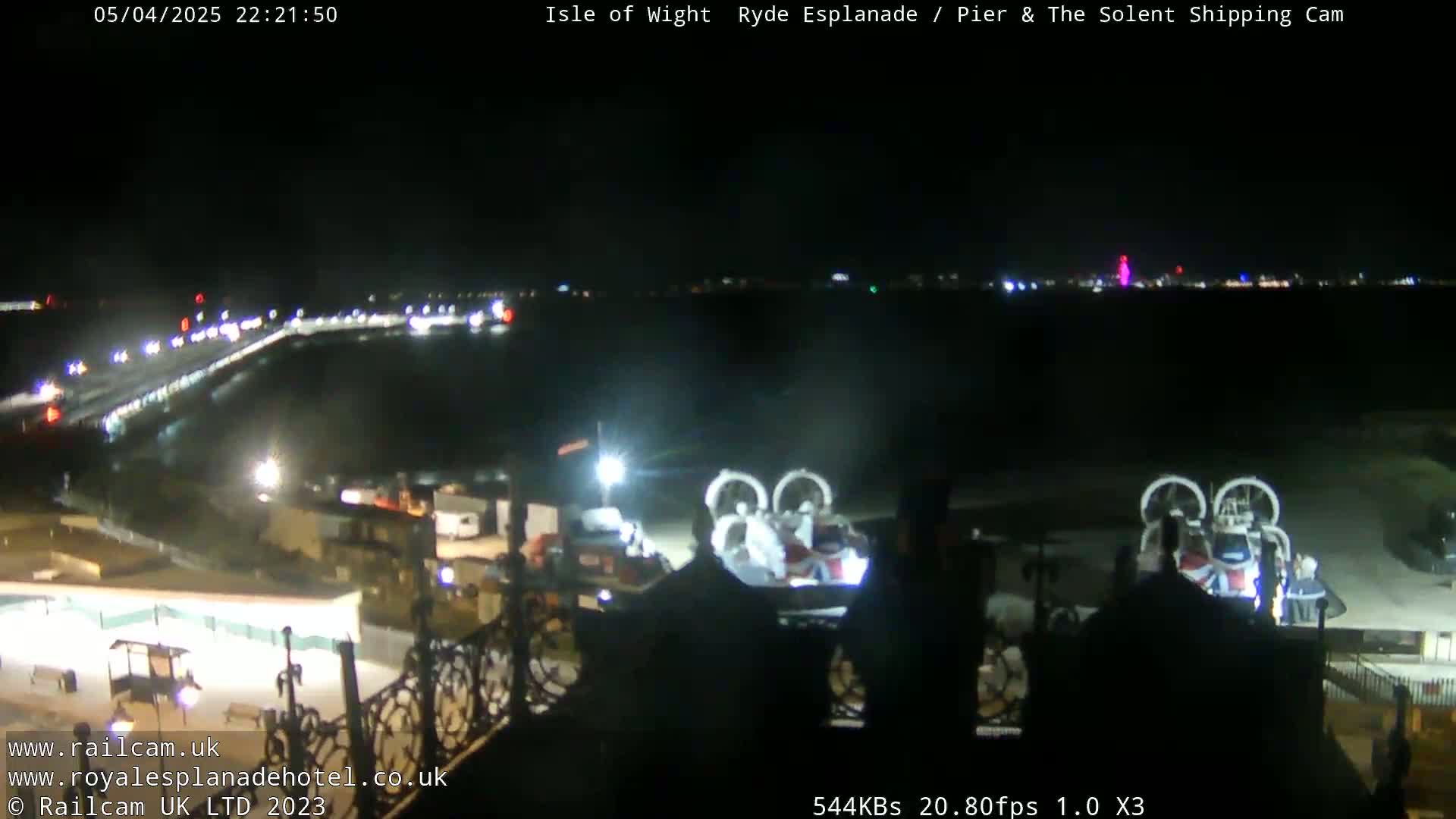 A nighttime view of a body of water with a hovercraft docked near a pier and city lights in the distance under a clear sky.