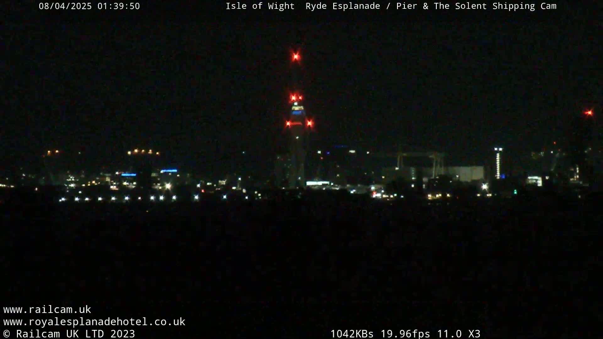A nighttime long shot of a city skyline with a tall structure illuminated with red lights, under a clear, dark sky.