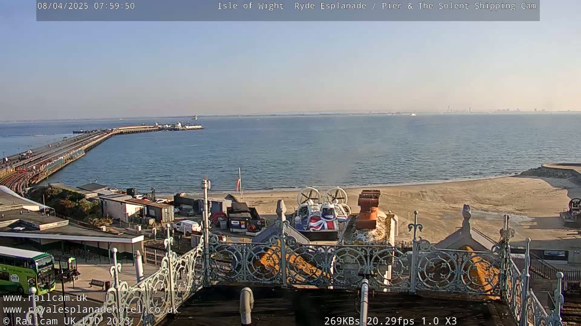 A hovercraft sits on a sandy beach near a pier extending into a calm, clear body of water under a bright, sunny sky.