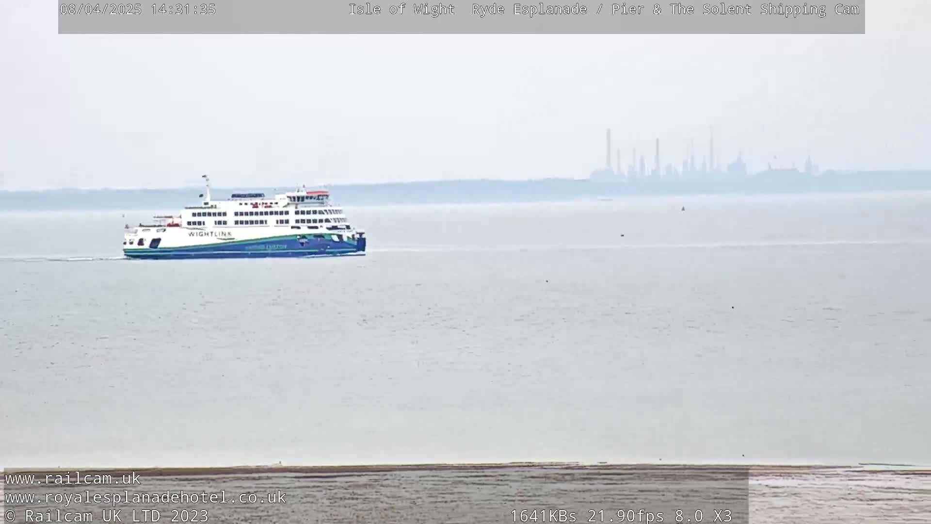 A ferry sails on a calm, hazy body of water with a distant industrial skyline visible on the horizon.