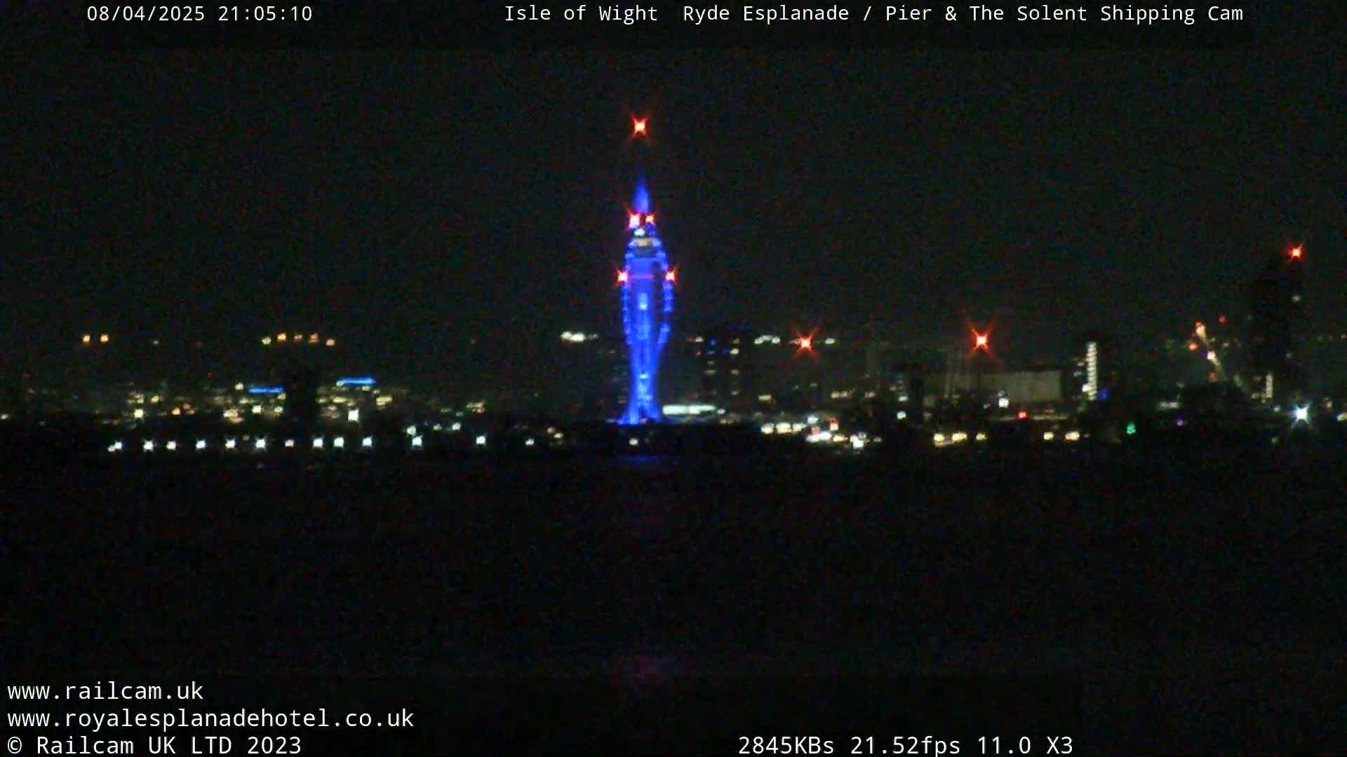 A nighttime, long shot of a city skyline across a body of water, featuring a tall structure illuminated in blue light.