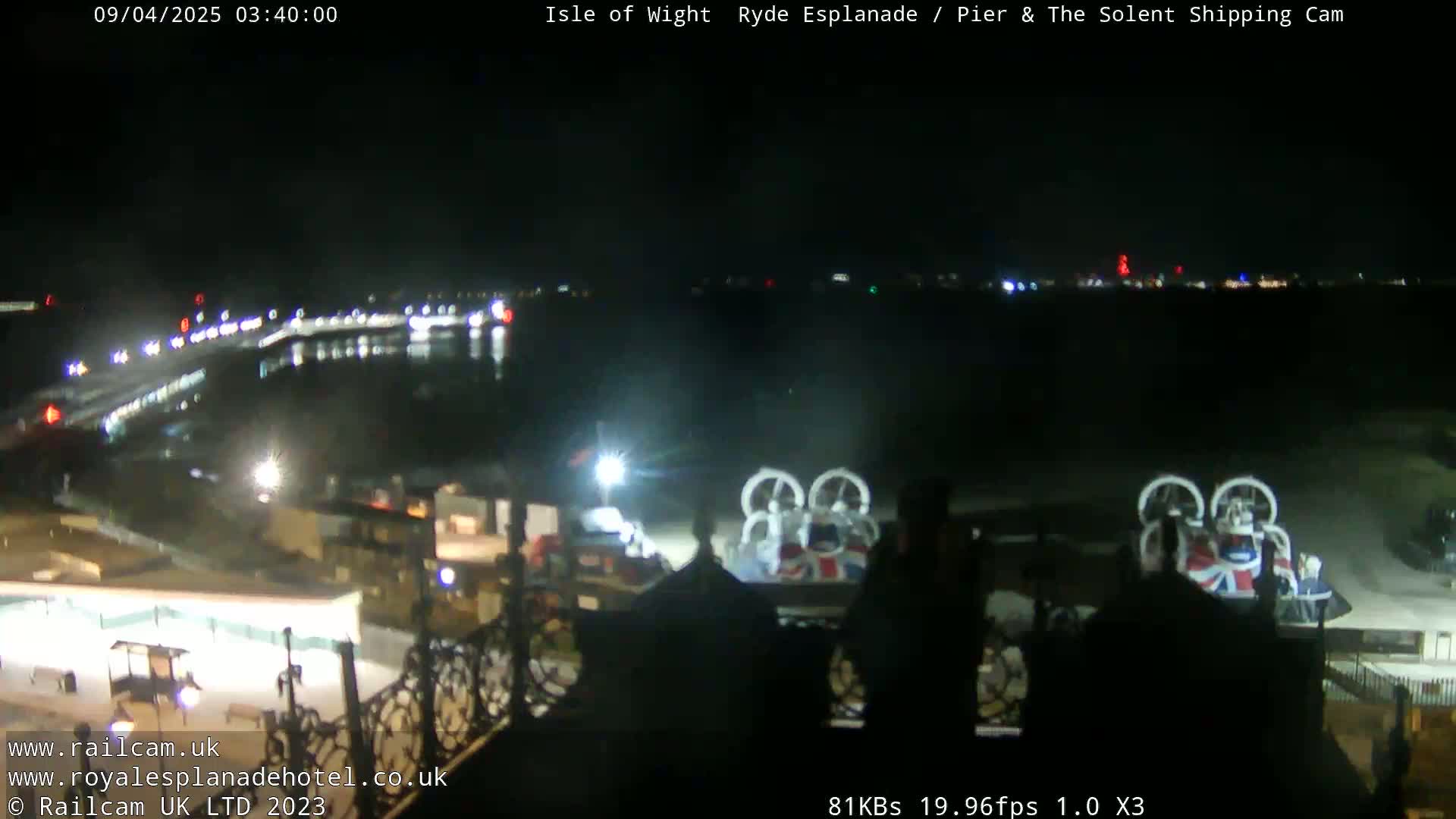 A nighttime view of a body of water with lights reflecting on it, a shoreline with buildings and a hovercraft parked near a pier.