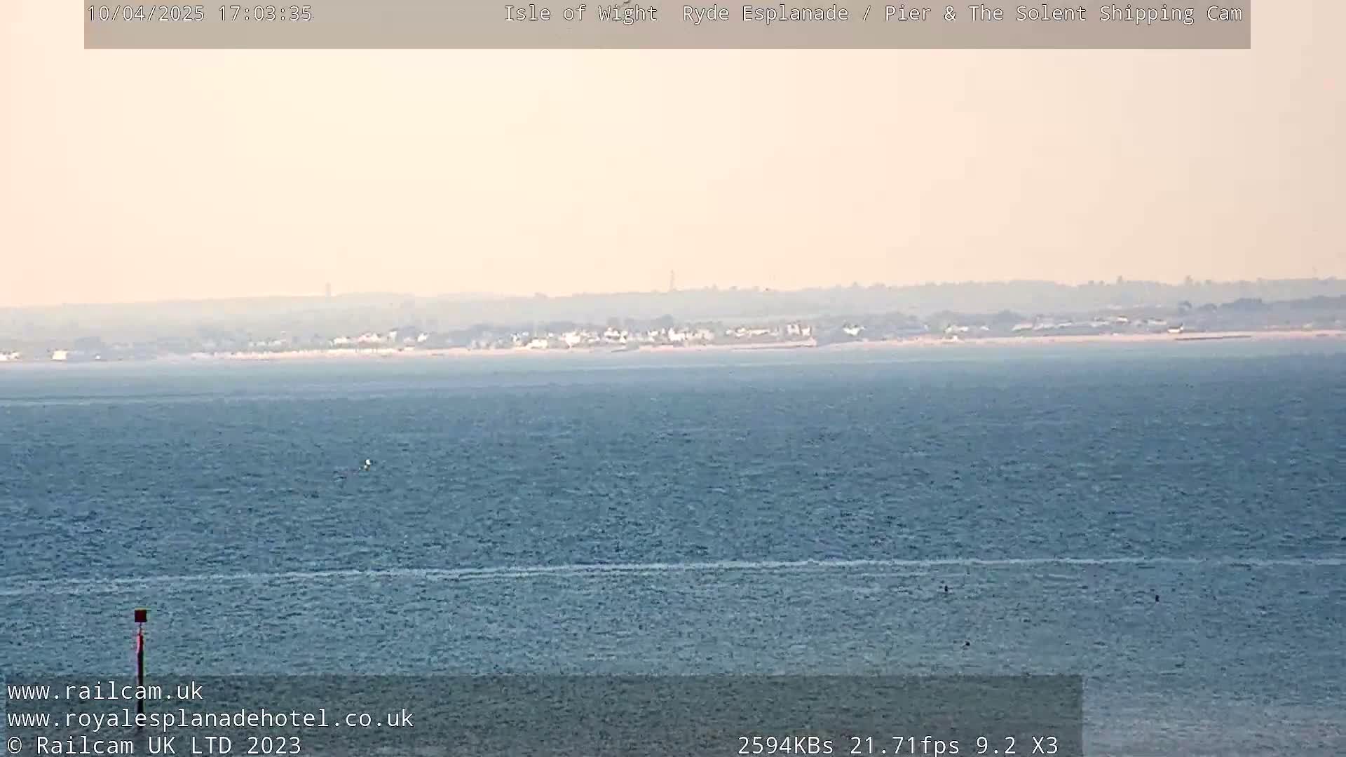 A hazy, long shot shows a calm body of water with a distant shoreline under a light, overcast sky.