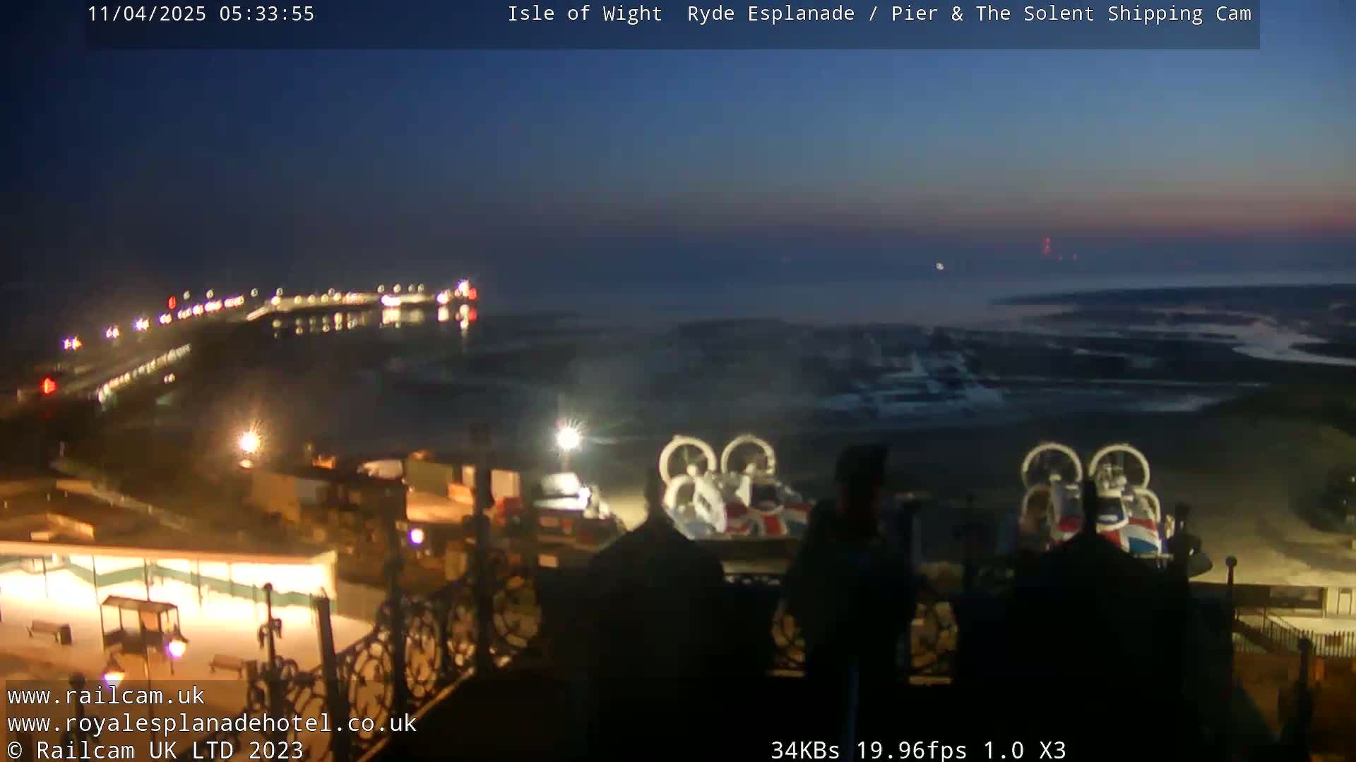 A nighttime, coastal scene shows a pier with lights, a body of water, and hovercrafts near a shoreline under a dark blue, twilight sky.