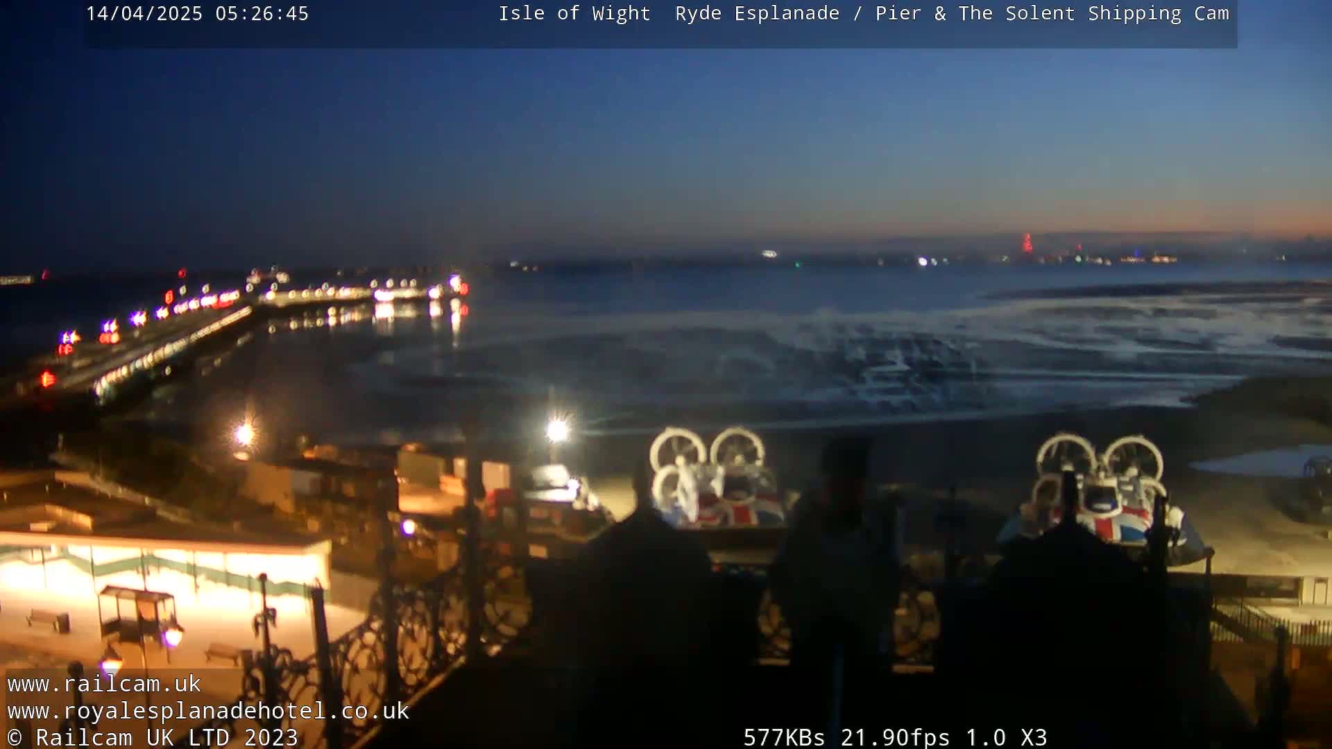 A nighttime view of a pier and shoreline, with a hovercraft parked on the beach and the dark, calm water reflecting lights in the distance.
