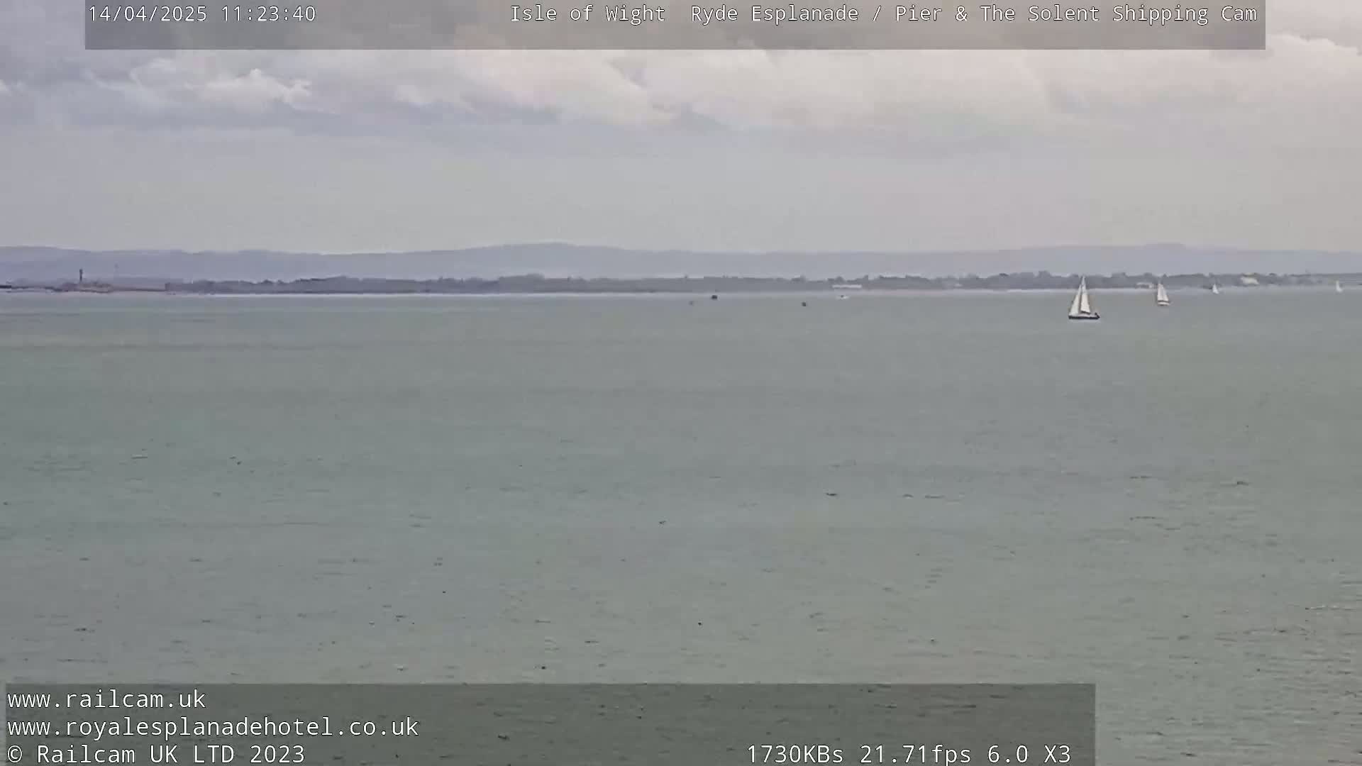A calm, overcast day shows a wide expanse of gray-green water with a few sailboats in the distance and a low-lying landmass on the horizon.