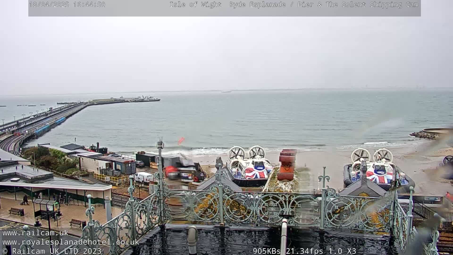 Two hovercrafts are on a beach near a pier and railway line, under an overcast sky.