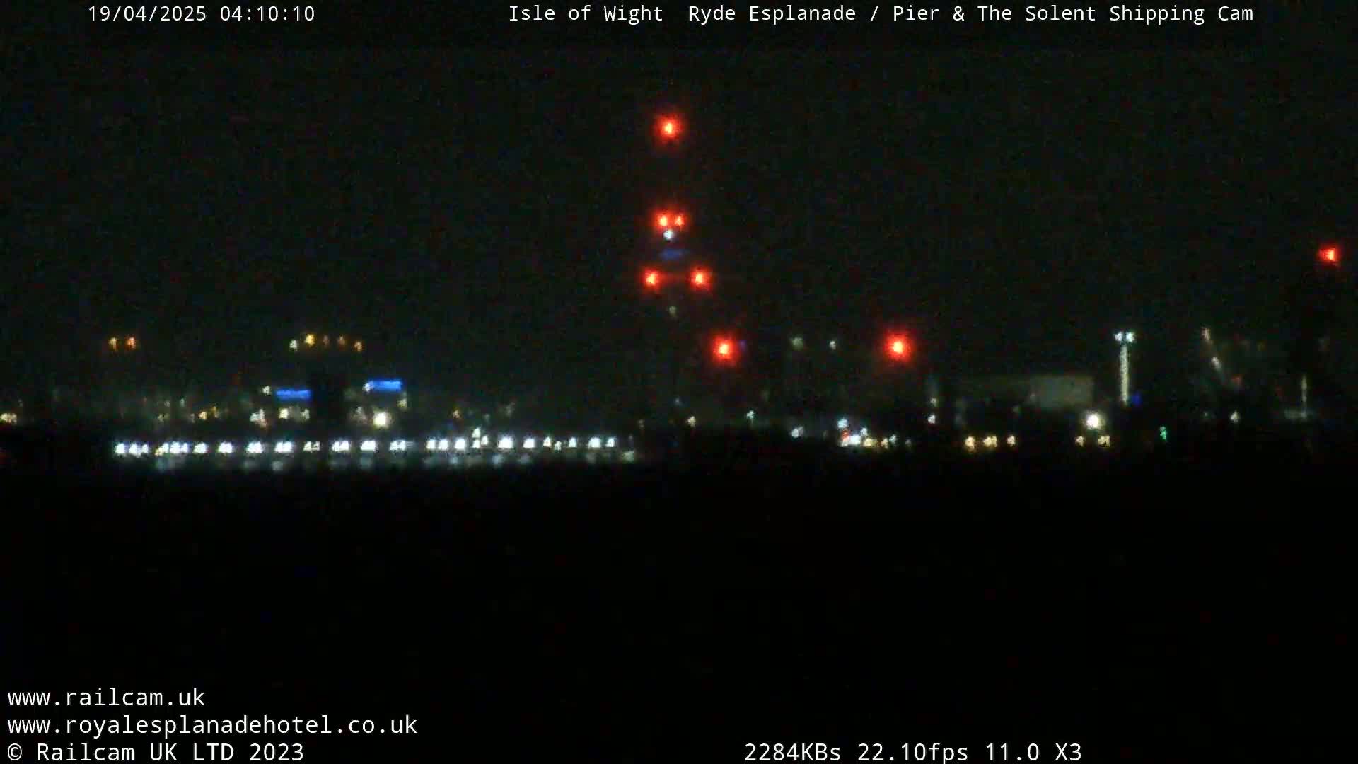 A nighttime long shot of a dark waterfront with scattered lights and a series of red lights ascending vertically in the distance.