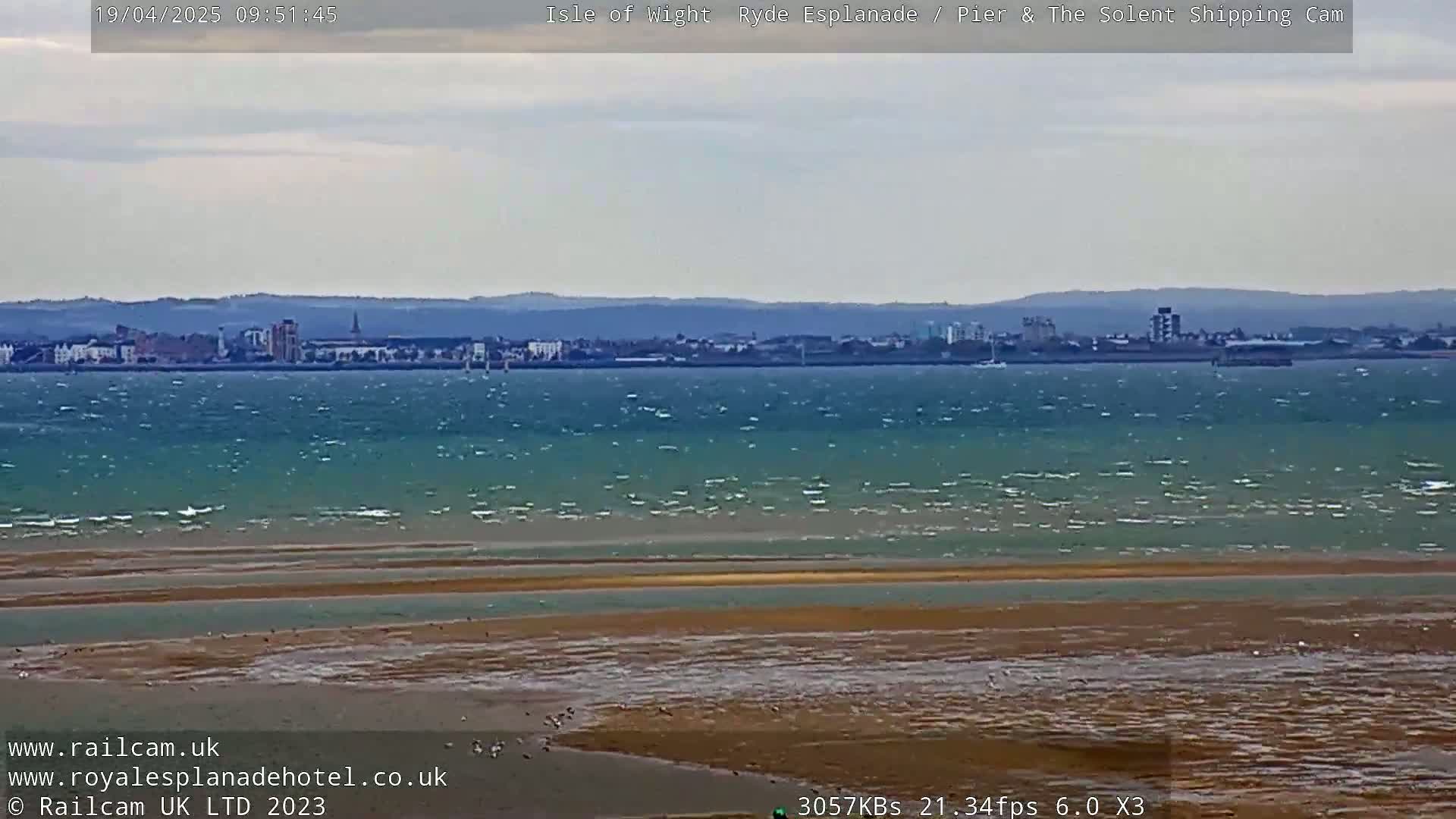 A windy day shows a wide expanse of choppy water separating a sandy shore from a distant cityscape.