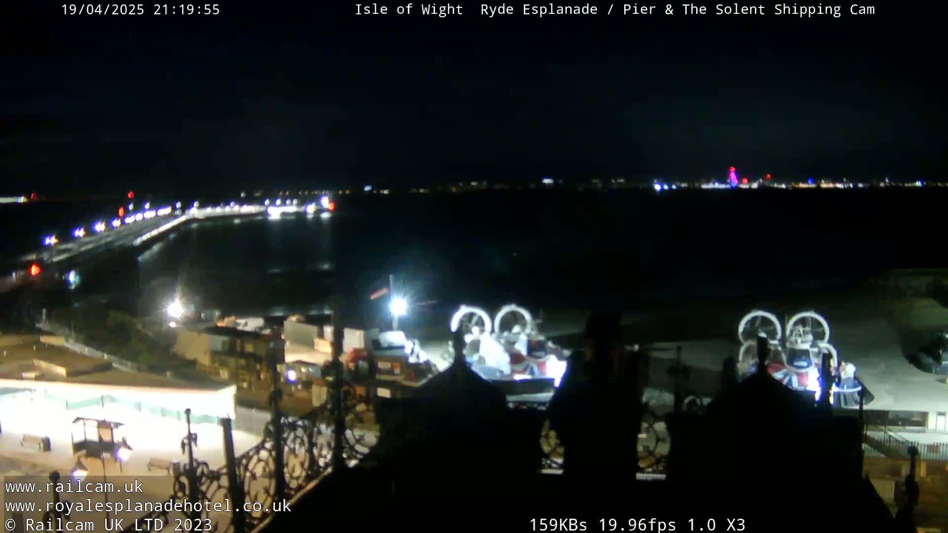 A nighttime view of a harbor with a pier, illuminated buildings, and a hovercraft, under a clear, dark sky.