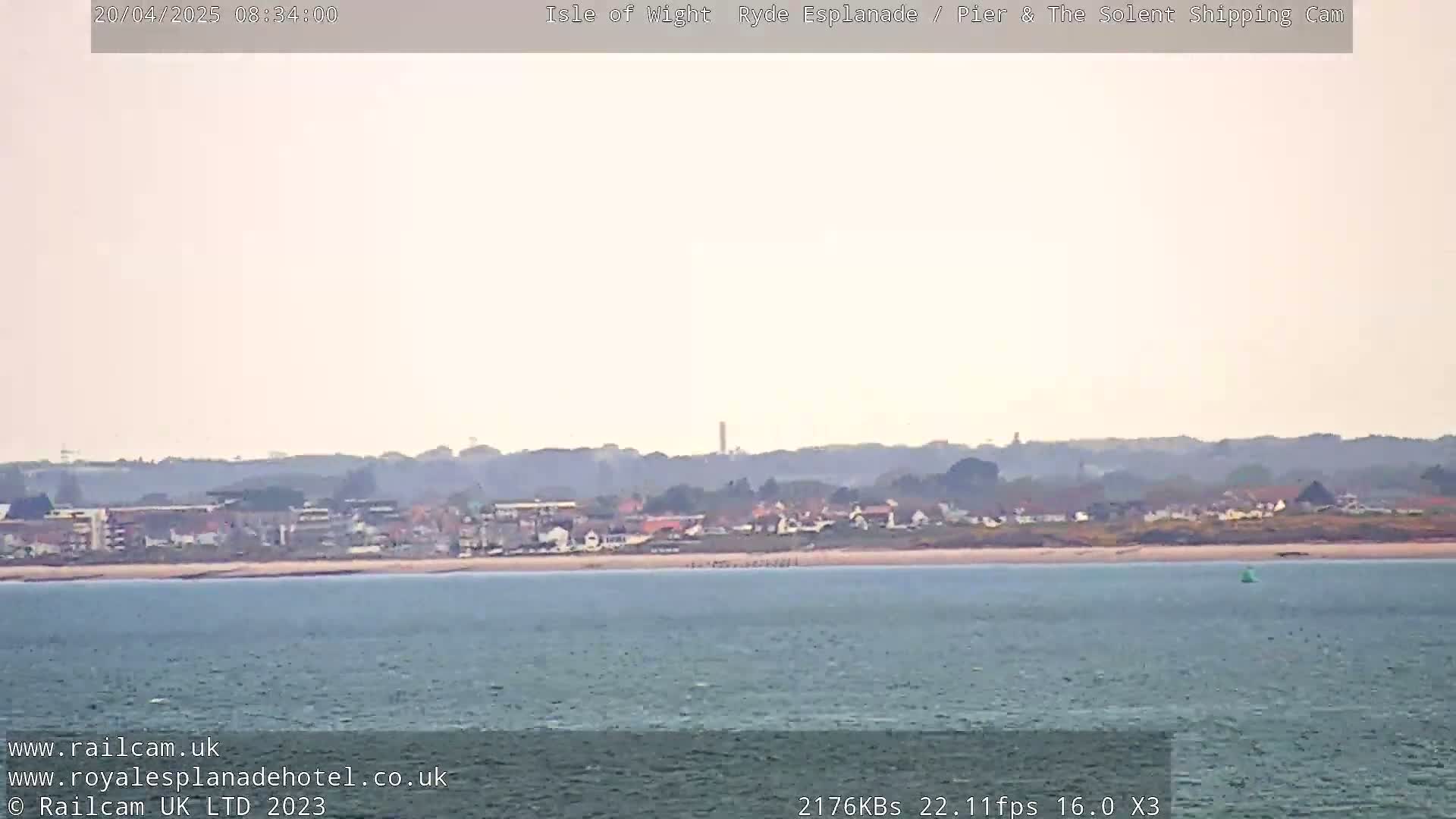 A calm body of water stretches out to a distant shoreline featuring a town and sandy beach under a hazy sky.