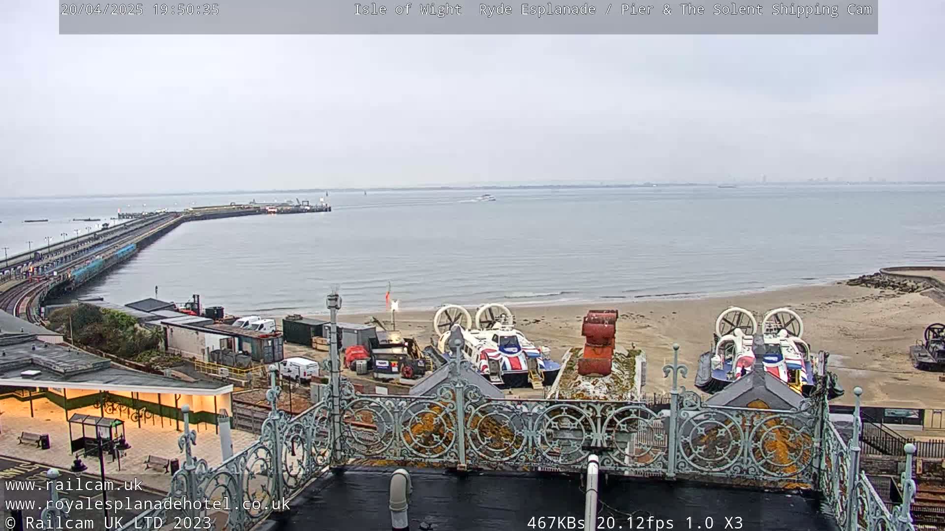 An overcast day shows a pier extending into a body of water, with hovercrafts on a sandy beach in the foreground and a railway line visible to the left.