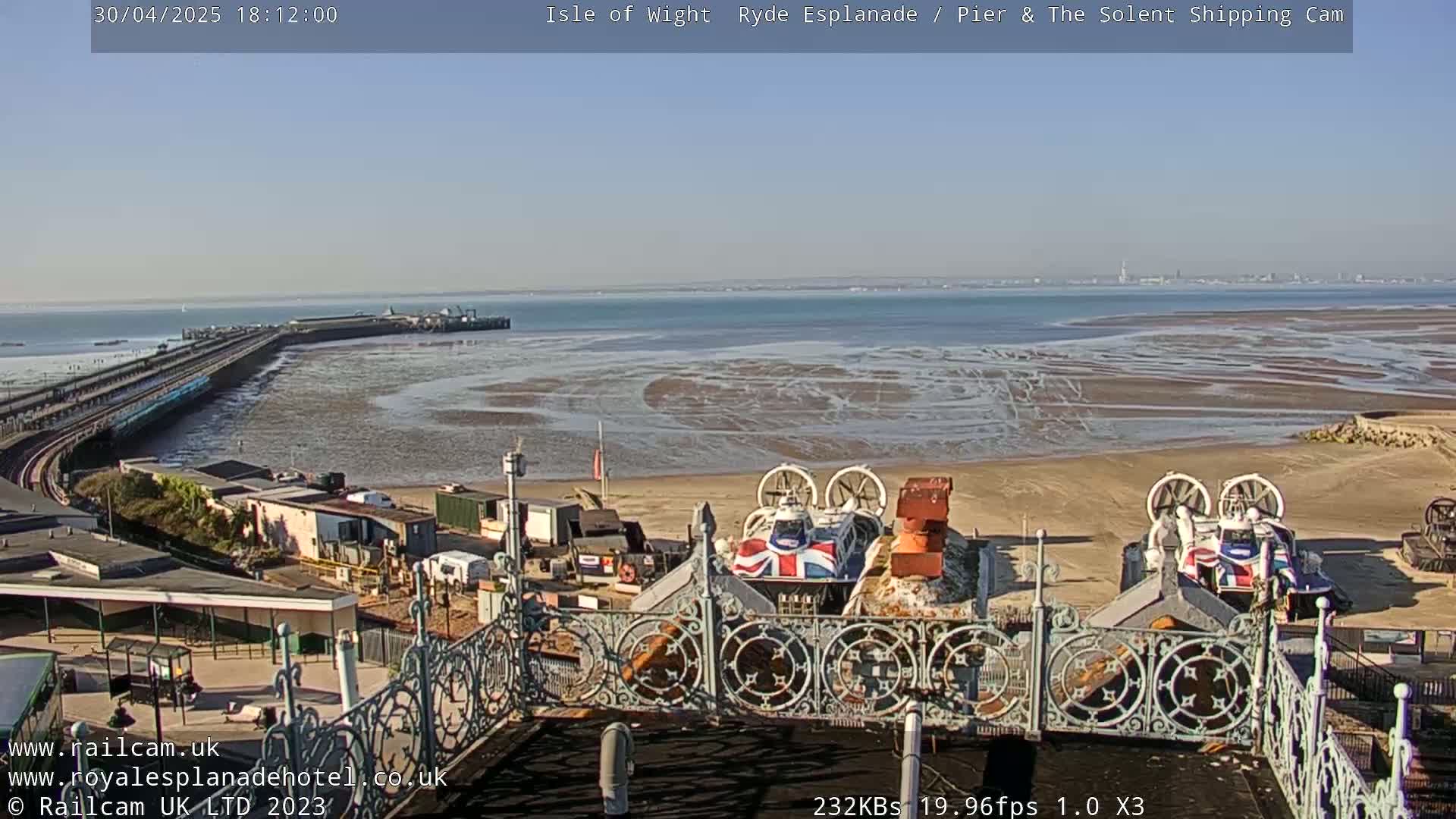 A sunny seaside scene shows a long pier extending into a wide expanse of shallow, sandy water, with two hovercrafts parked on the beach in the foreground and a metal railing in the immediate foreground.