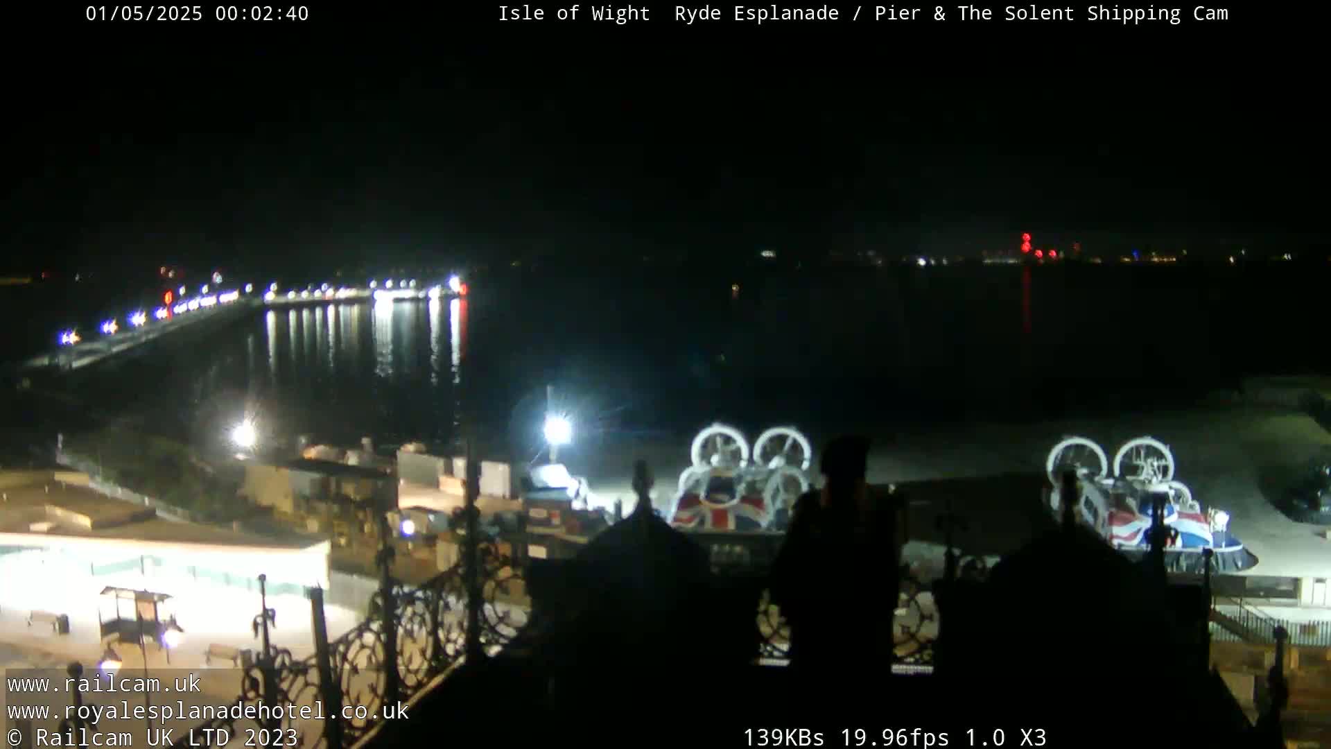 A nighttime view of a body of water with lights reflecting on its surface, a pier, and two hovercrafts parked on land.