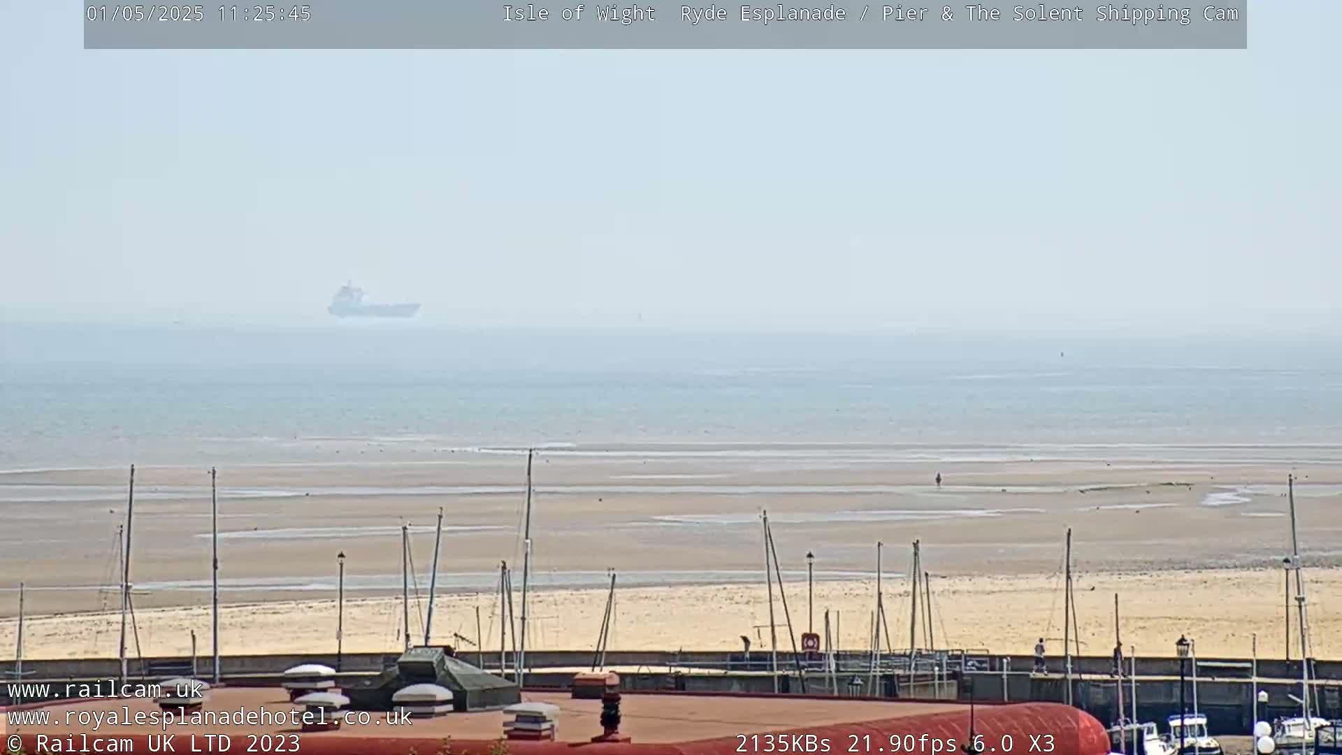 A hazy view of a sandy beach with sailboats, a distant ship on the ocean, and a low-lying building in the foreground.