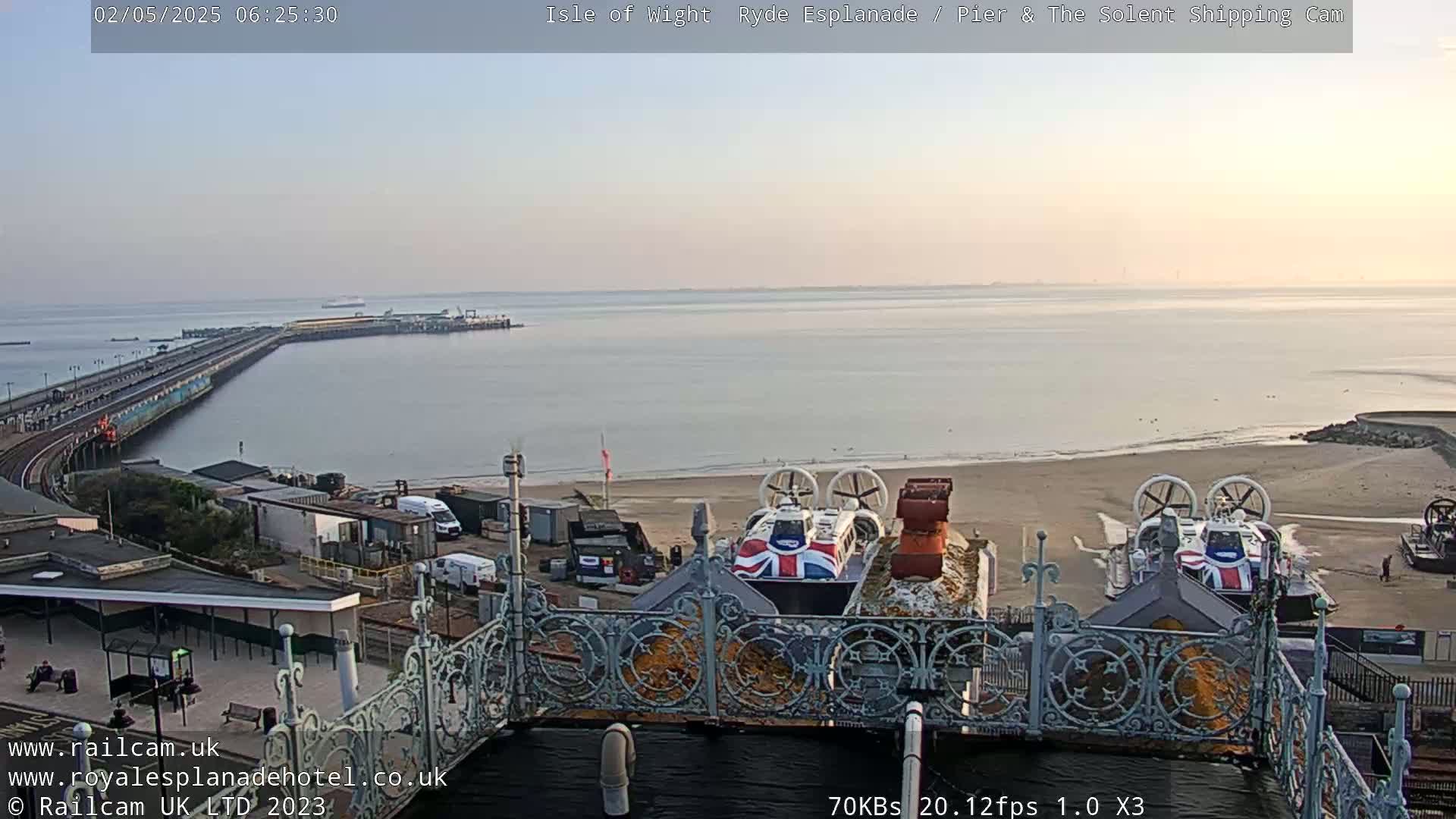 A calm, pale-blue body of water stretches to the horizon, featuring a long pier extending from the shore, where two hovercrafts rest near a decorative railing.