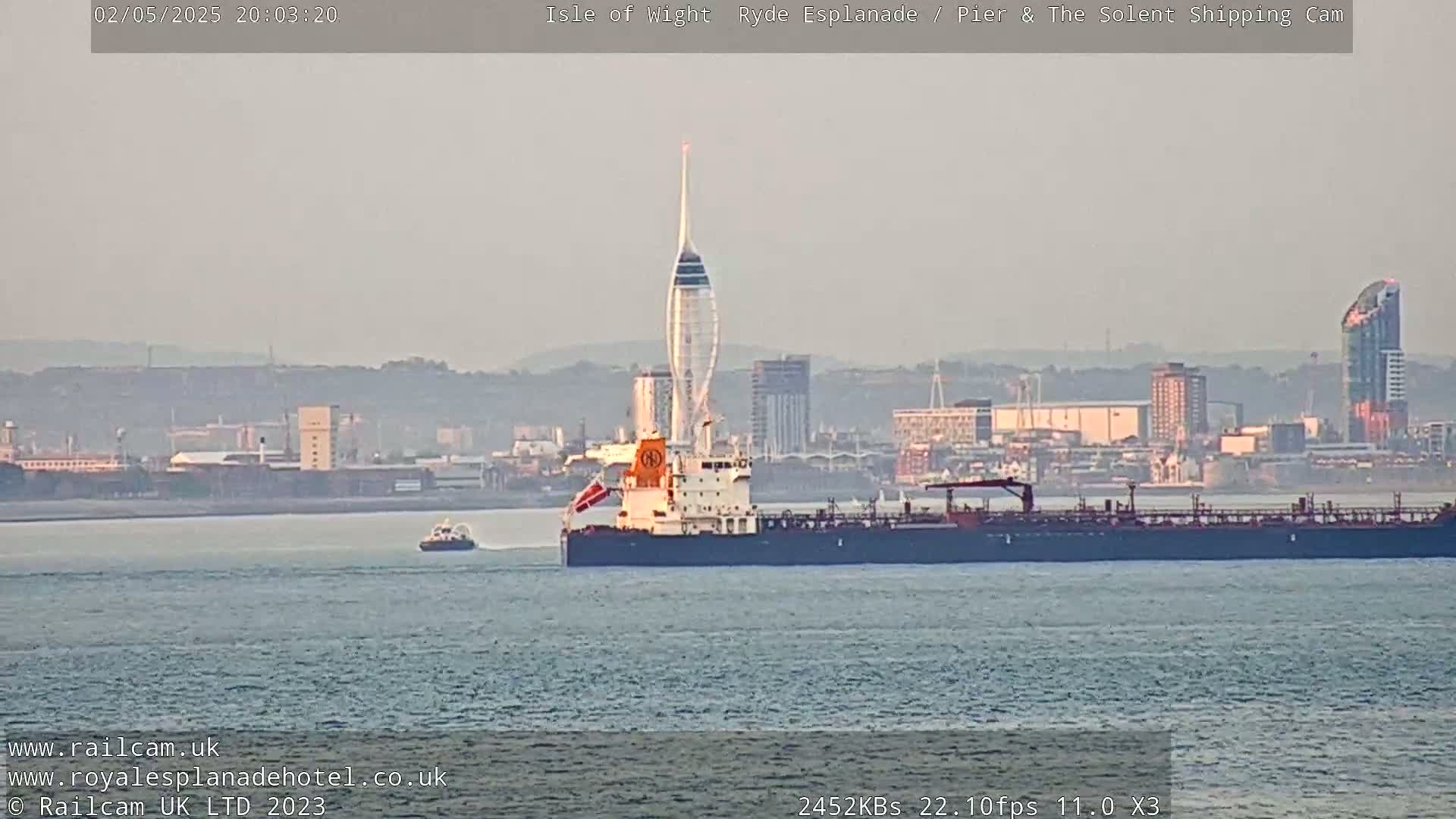 A large tanker and a smaller vessel are on calm water in front of a hazy cityscape with a tall, slender tower.