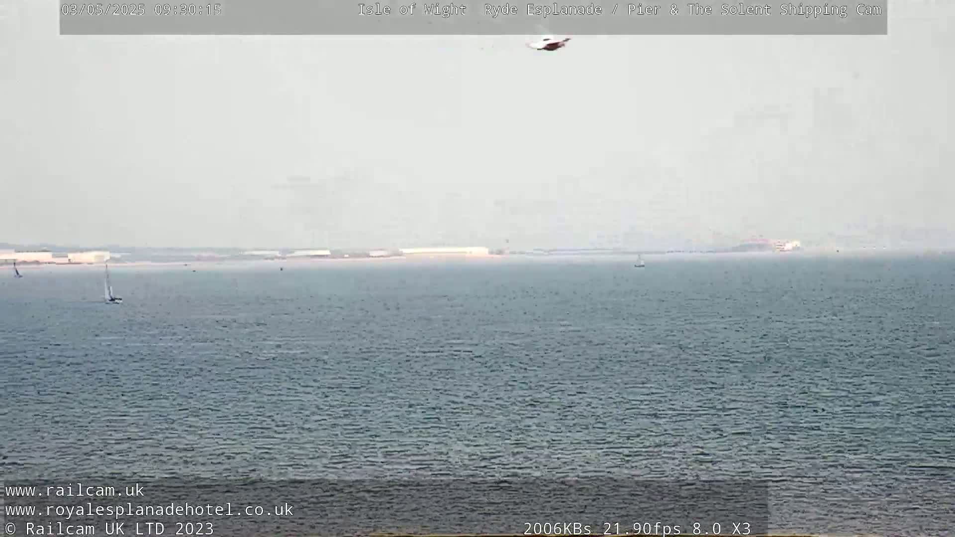 A helicopter is seen flying over a calm, hazy body of water with several small boats and a distant shoreline.