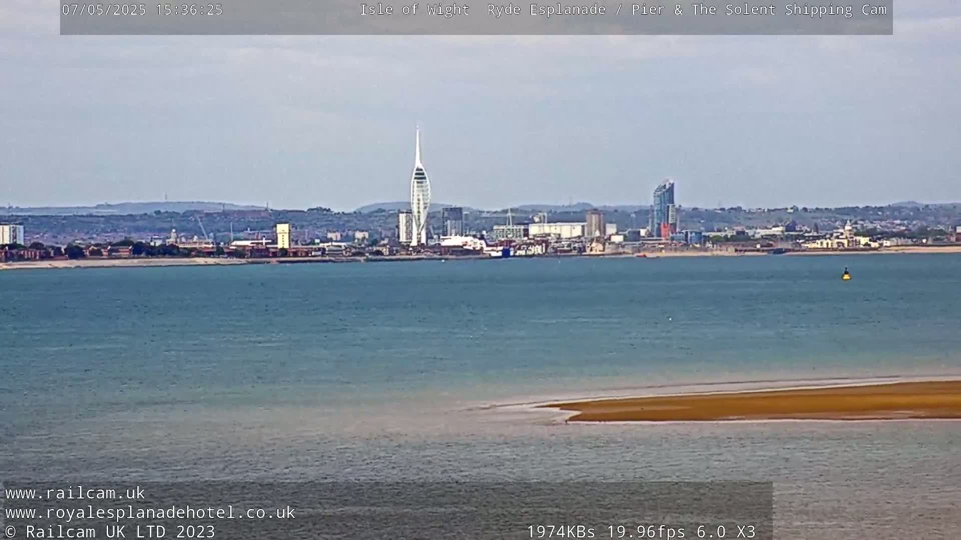 A long shot across calm, light-blue water shows a distant city skyline with a tall, slender tower under a mostly clear sky.