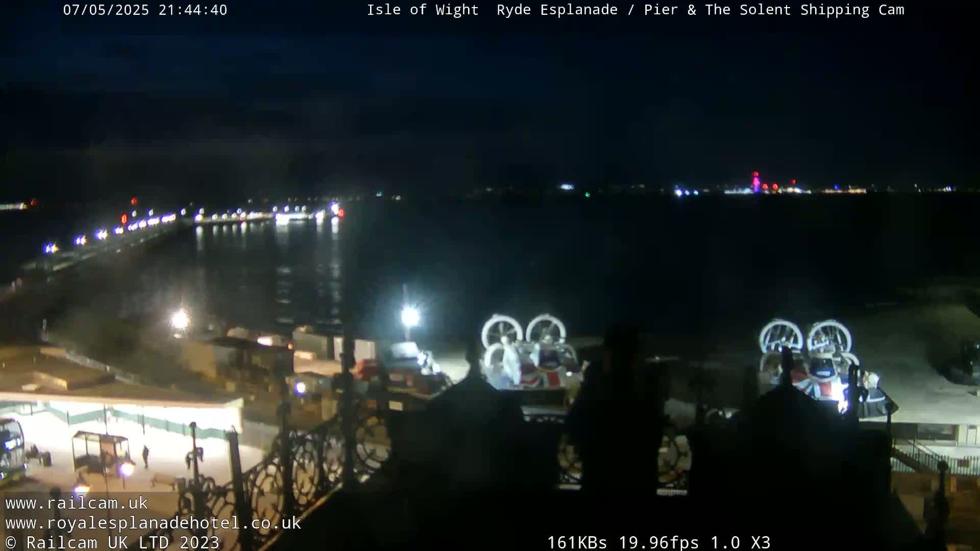 A nighttime view of a pier and body of water, with lights illuminating a roadway and several hovercrafts parked nearby.