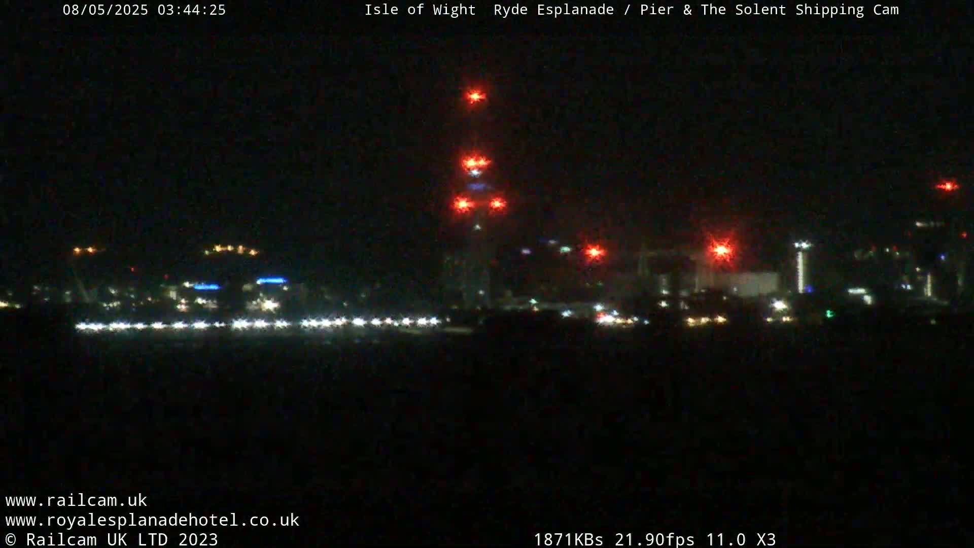 A nighttime, dark scene shows a distant cityscape with lights, illuminated by several bright red lights on a tall structure.