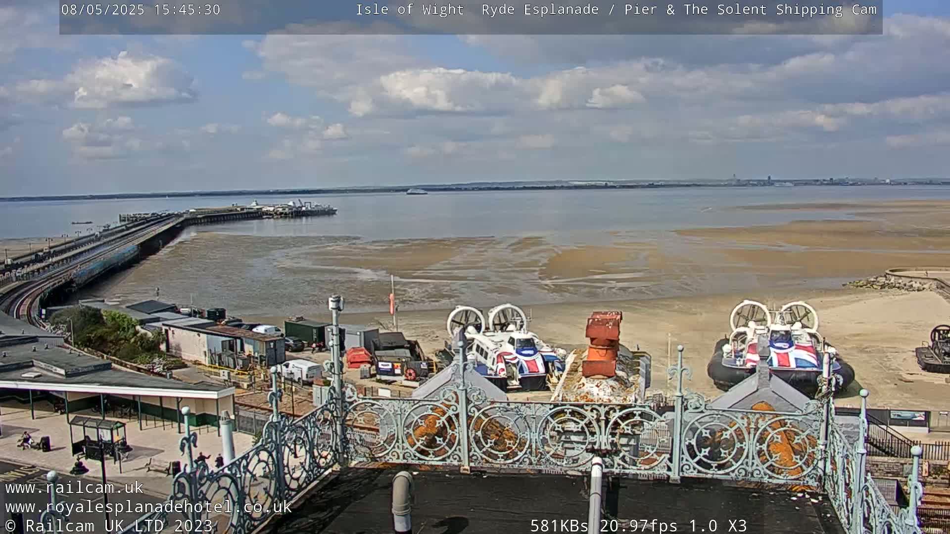 Two hovercrafts are parked on a beach near a pier under a partly cloudy sky.
