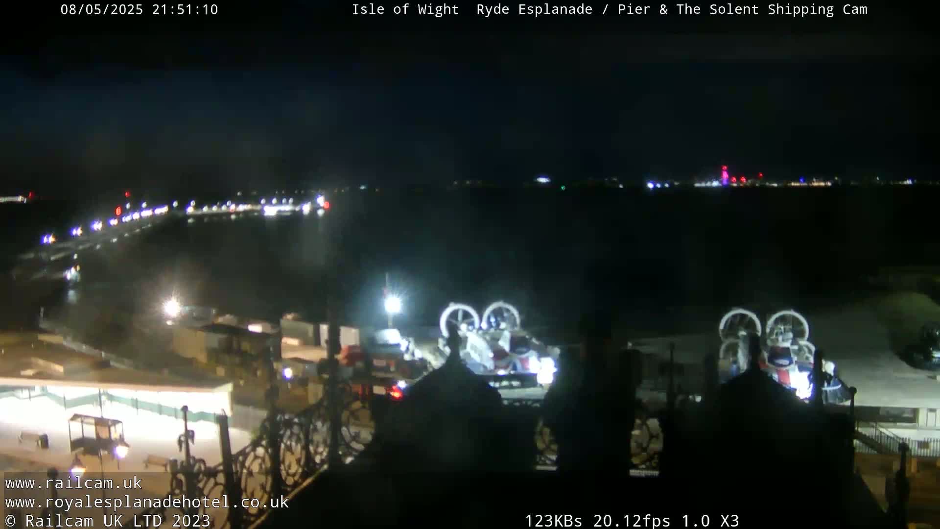 At night, a body of water is visible with lights in the distance, and two hovercrafts are docked near a structure.