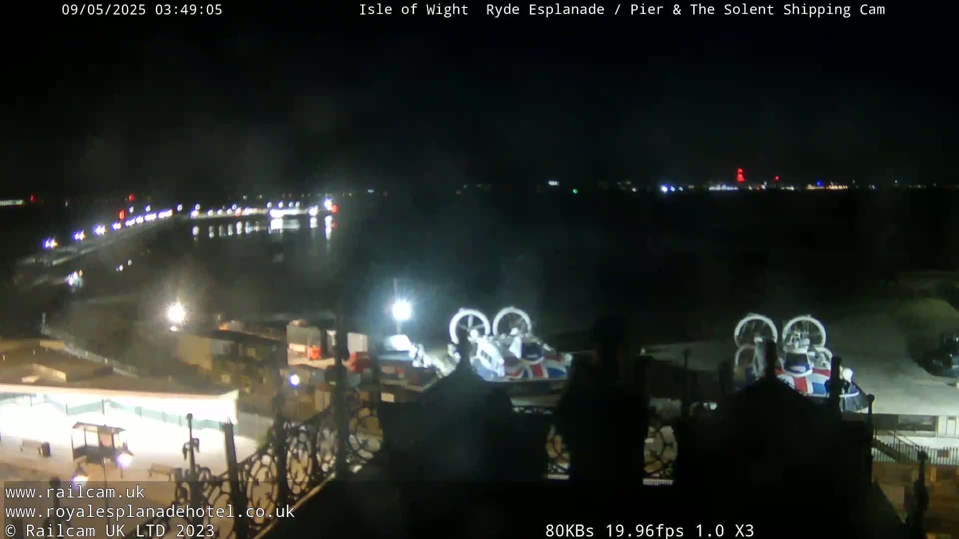 A nighttime view of a harbor with docked hovercrafts, traffic on a road, and distant lights across the water.