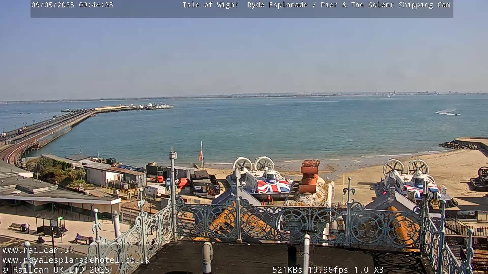A sunny, seaside scene shows a long pier extending into calm blue water, with two hovercrafts on a sandy beach in the foreground, and distant land visible across the water.